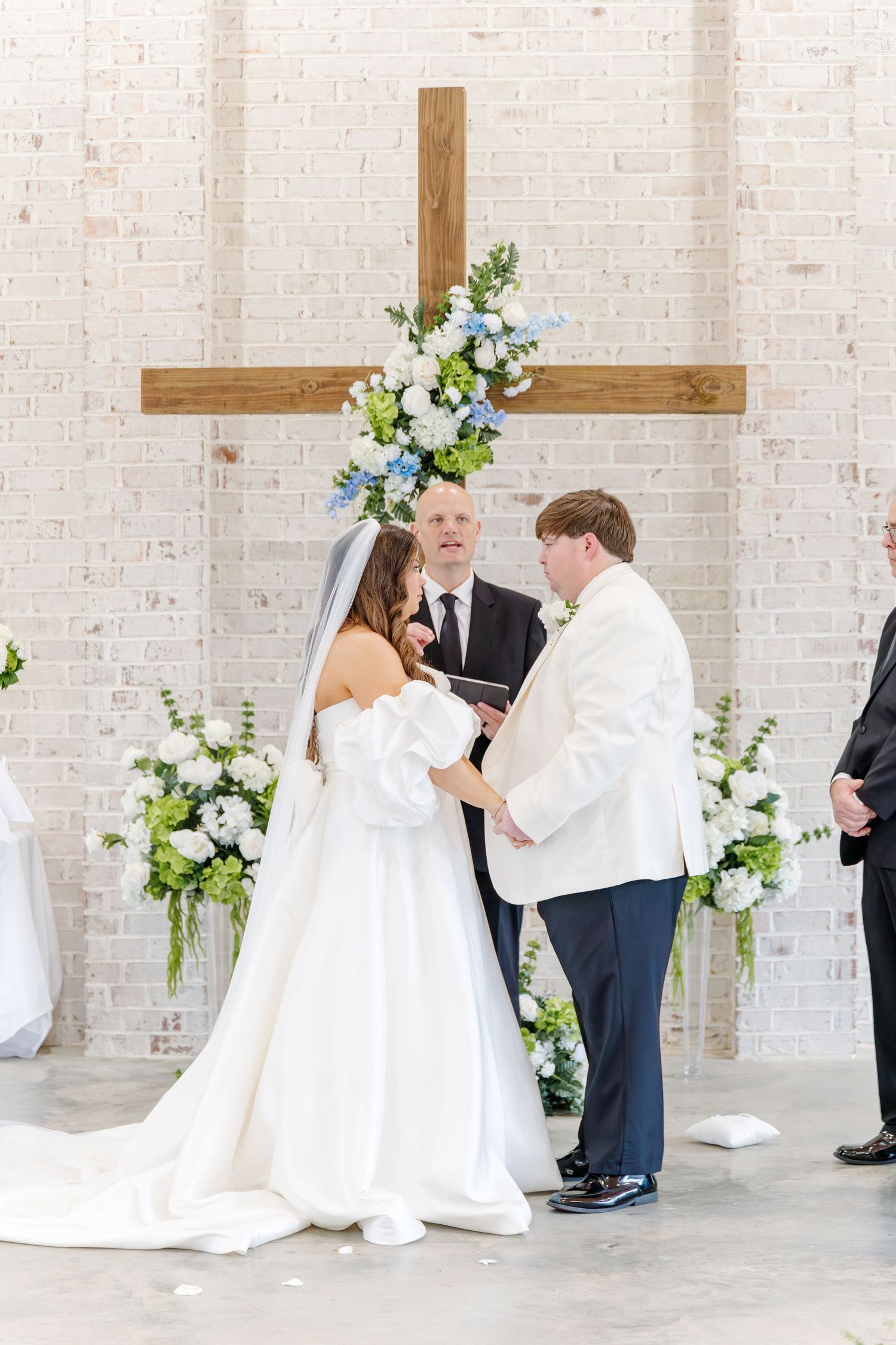 Bride and groom exchange vows before a wooden cross adorned with blue and white floral arrangements.