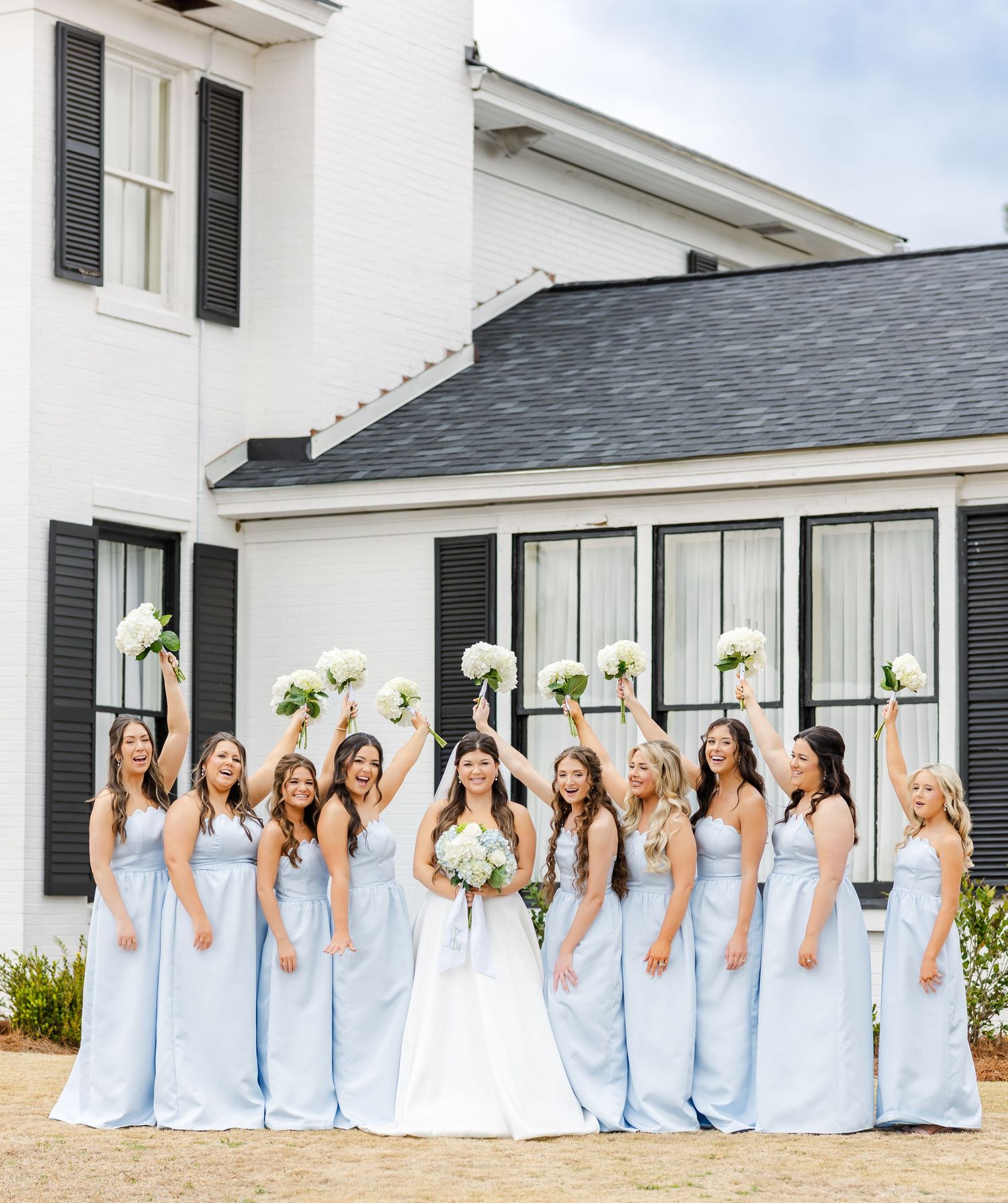 Bride with bridesmaids in light blue gowns raising bouquets of white flowers outside a white venue.