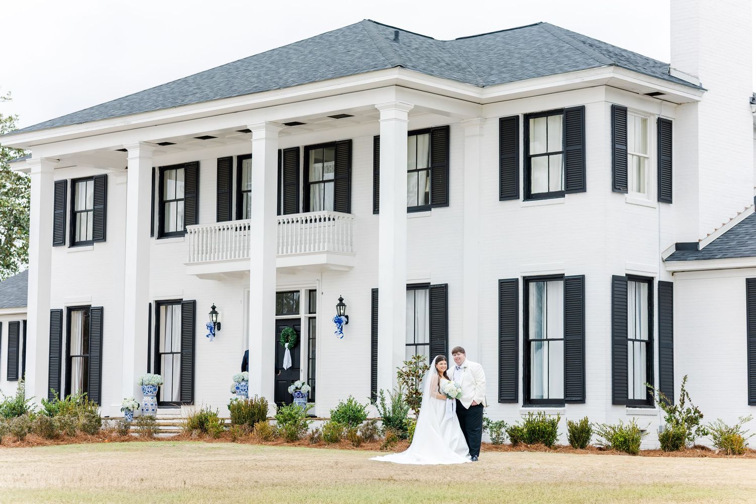 Bride and groom pose in front of elegant white Southern colonial wedding venue with black shutters.