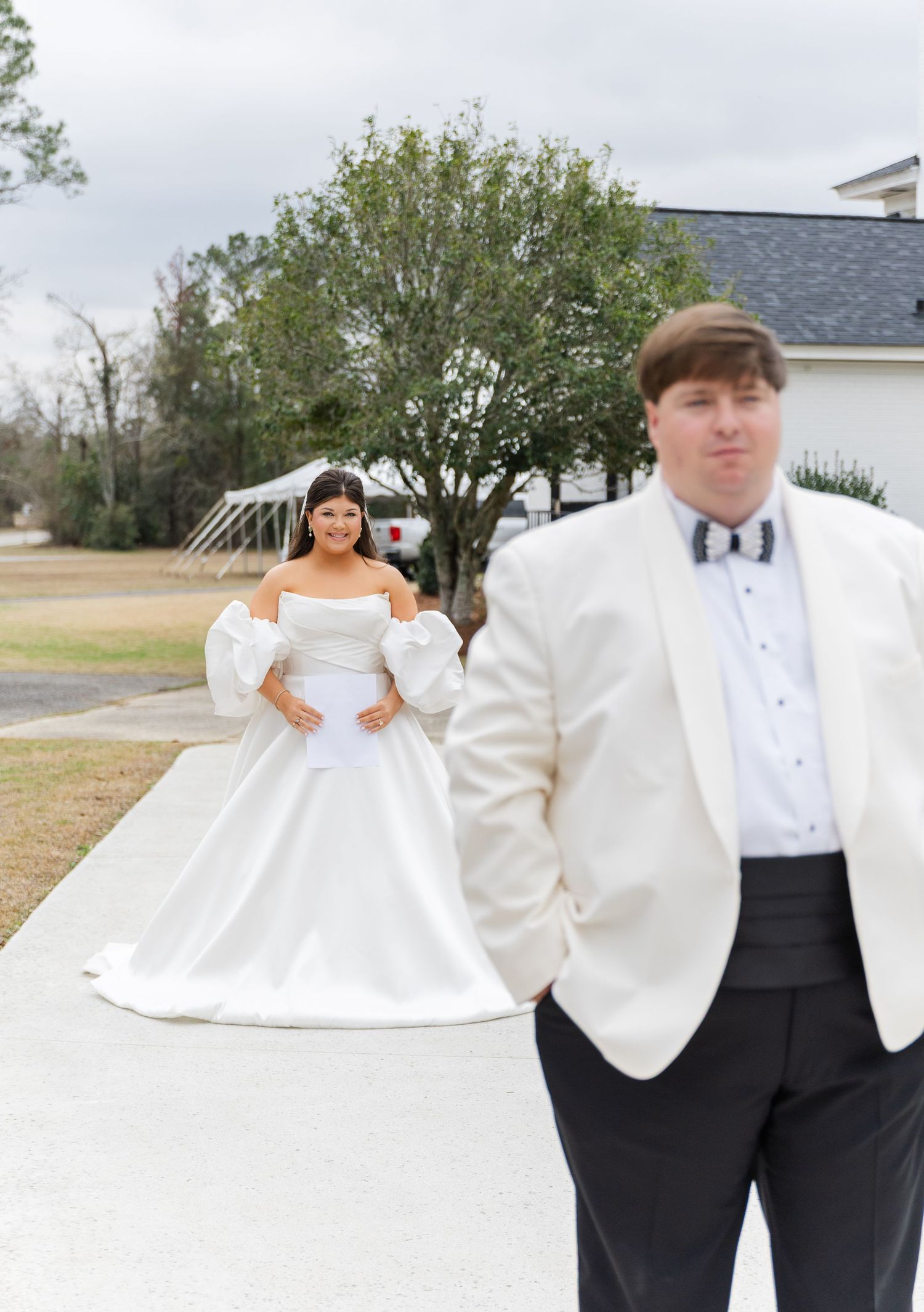 Bride in off-shoulder ballgown with puff sleeves stands behind groom in white tuxedo jacket on wedding day.