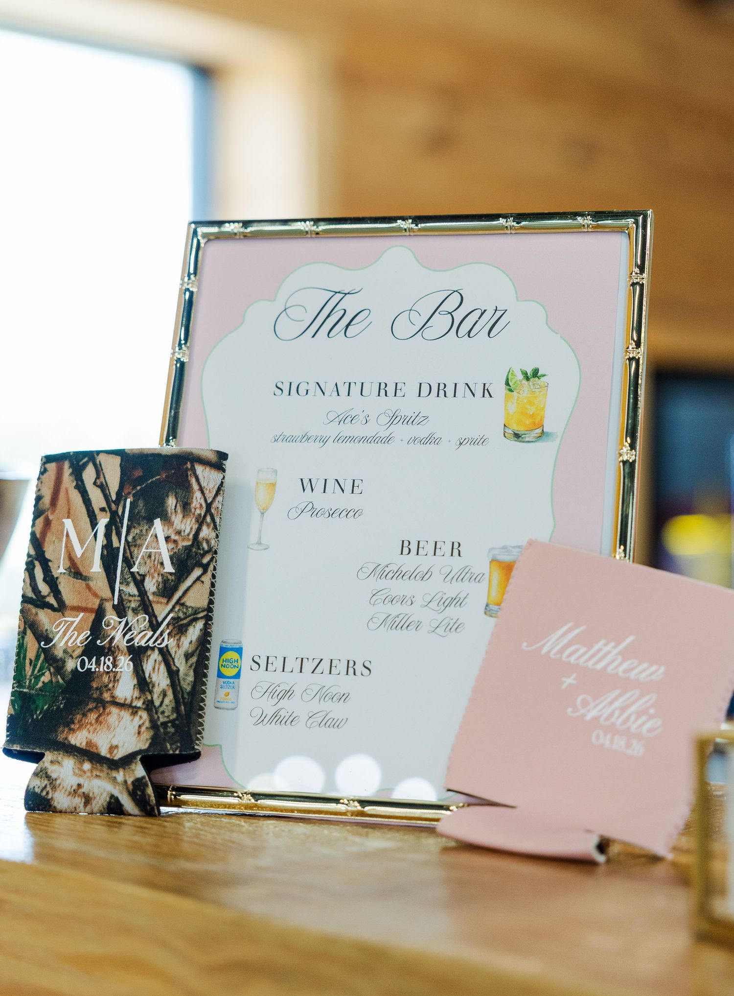 Wedding bar menu sign in gold frame with signature drinks list, beside camo and pink personalized koozies on wooden table.