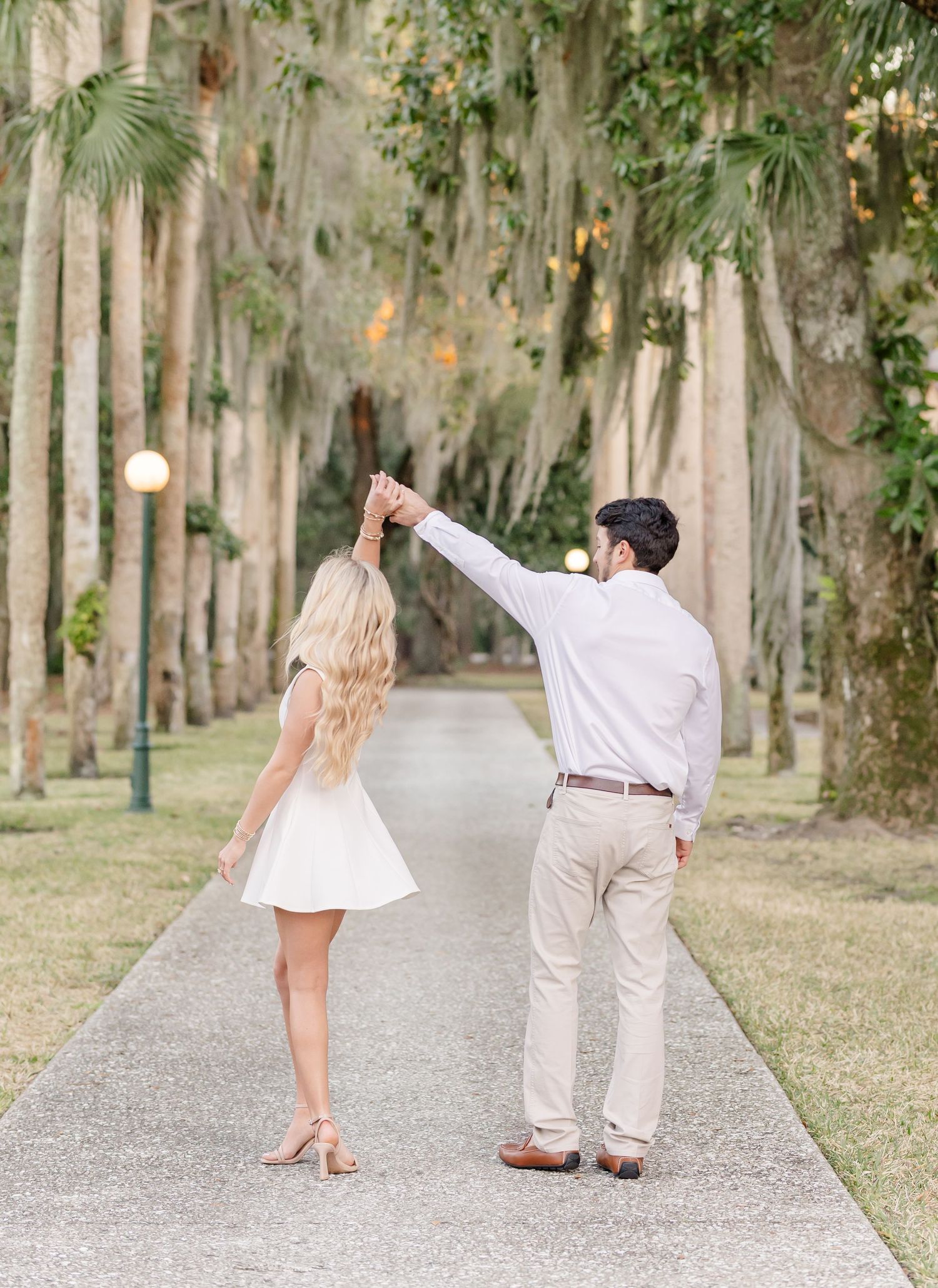 A couple dances on a tree-lined pathway, man spinning woman in white dress during outdoor engagement photo session.