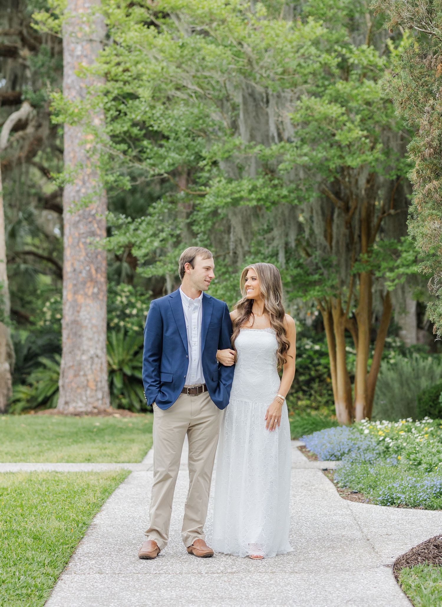Couple poses on garden path, man in navy blazer and woman in light blue gown, surrounded by lush trees.