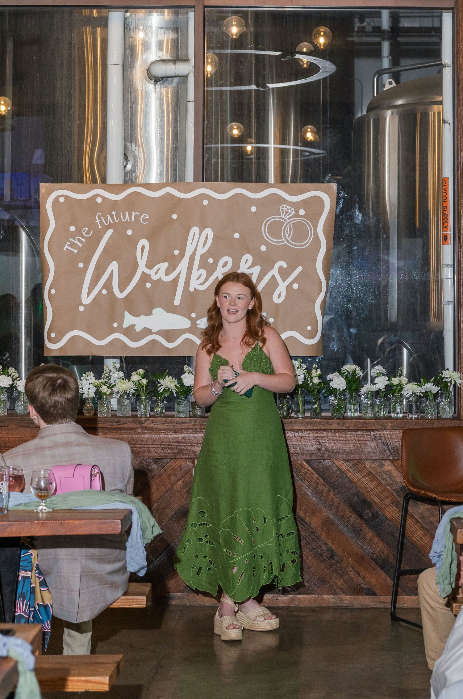 Woman in green dress gives a speech at The Future Walkers engagement party in a rustic brewery venue.