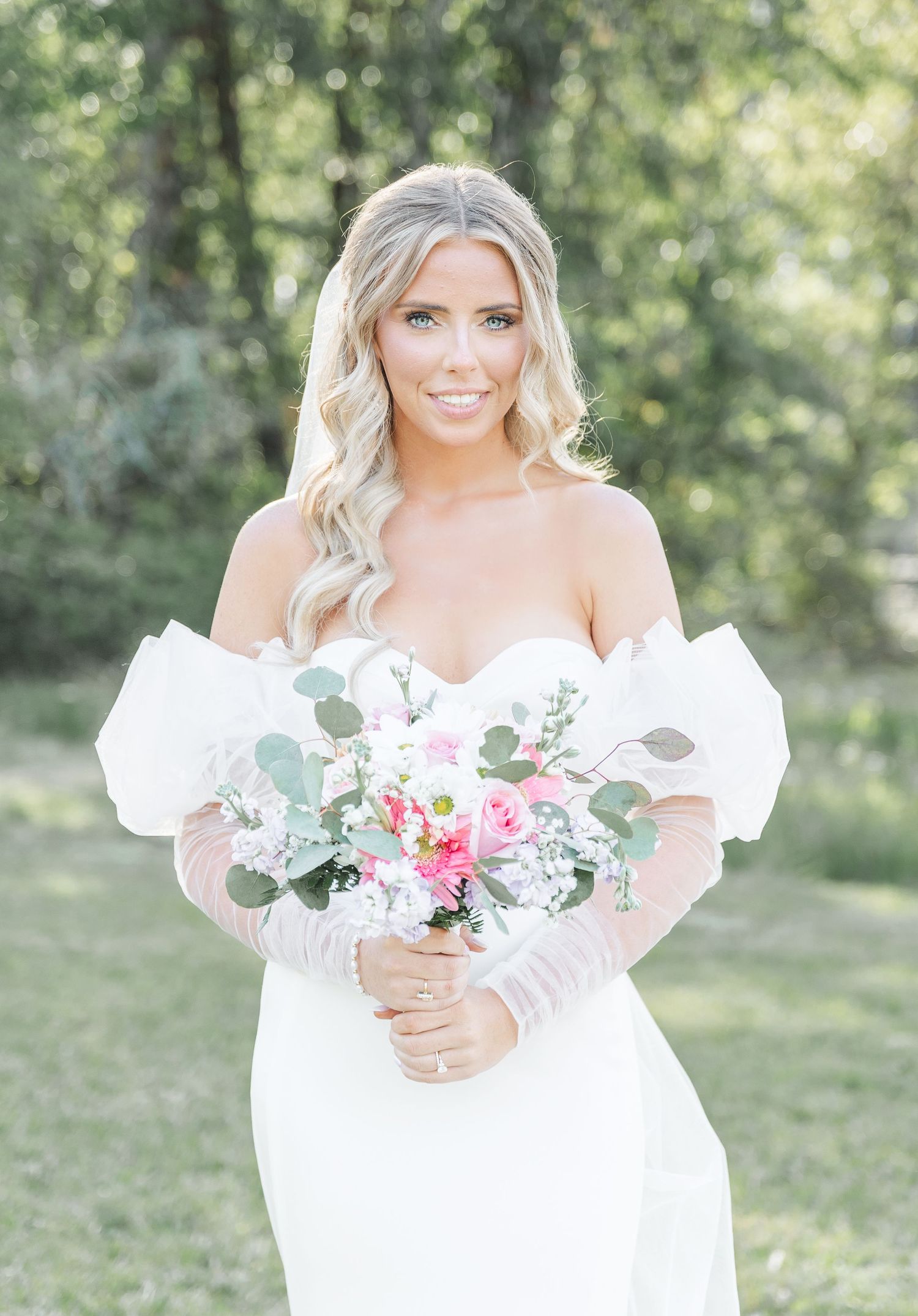 Smiling bride in elegant white dress poses with pink and white floral bouquet against green foliage.