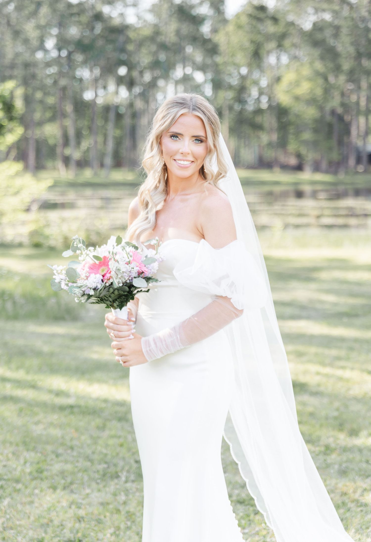 Elegant bride in a sleek white dress and veil holds a pink and white bouquet in a garden setting.