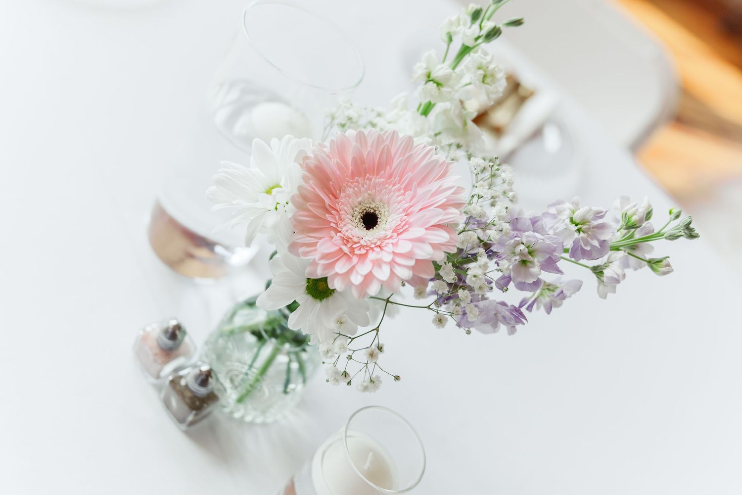 Soft focus image of pink gerbera daisy and purple flower arrangement on white table setting.