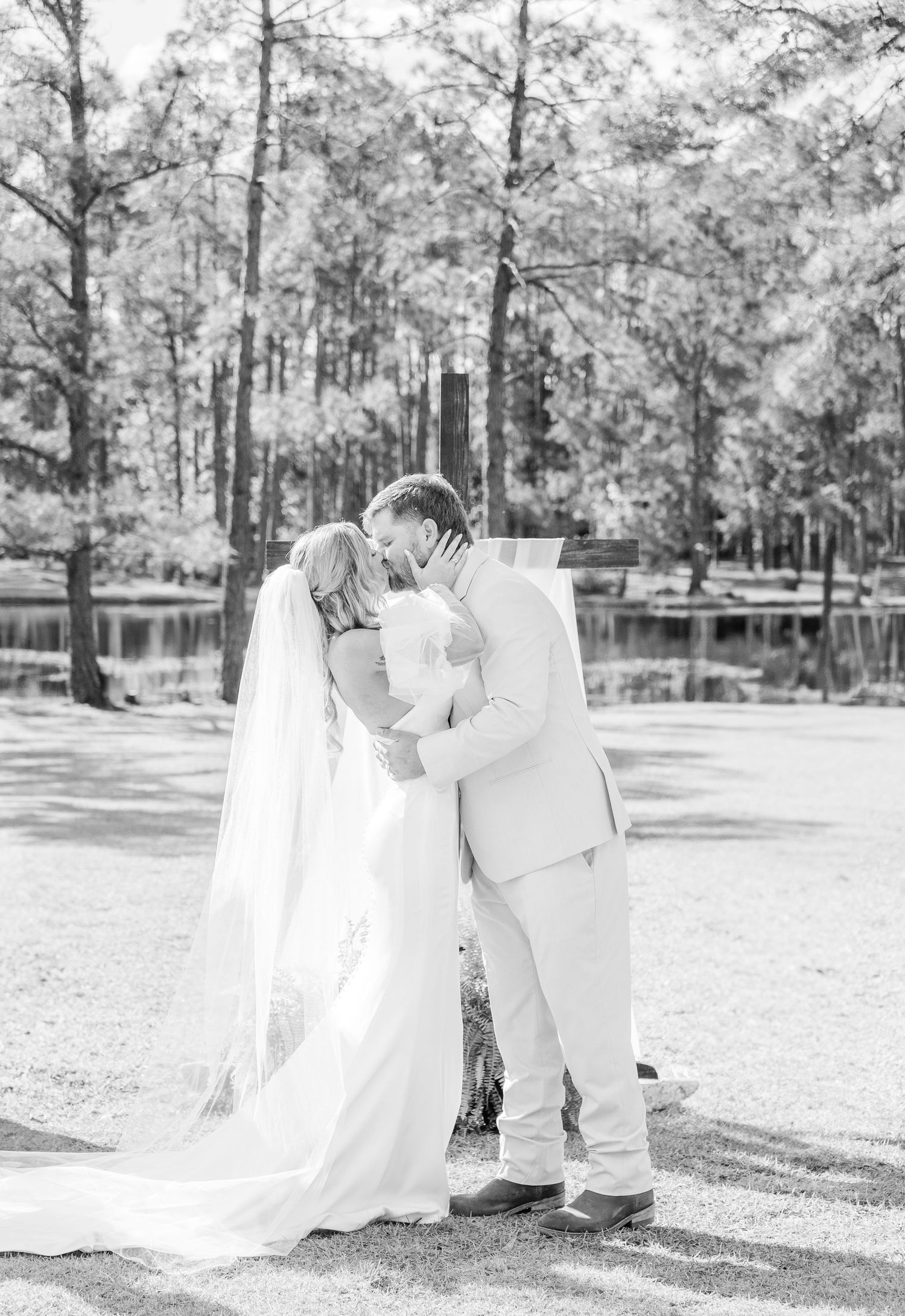 Romantic black and white wedding photo of a couple embracing by a serene lake surrounded by tall pine trees.