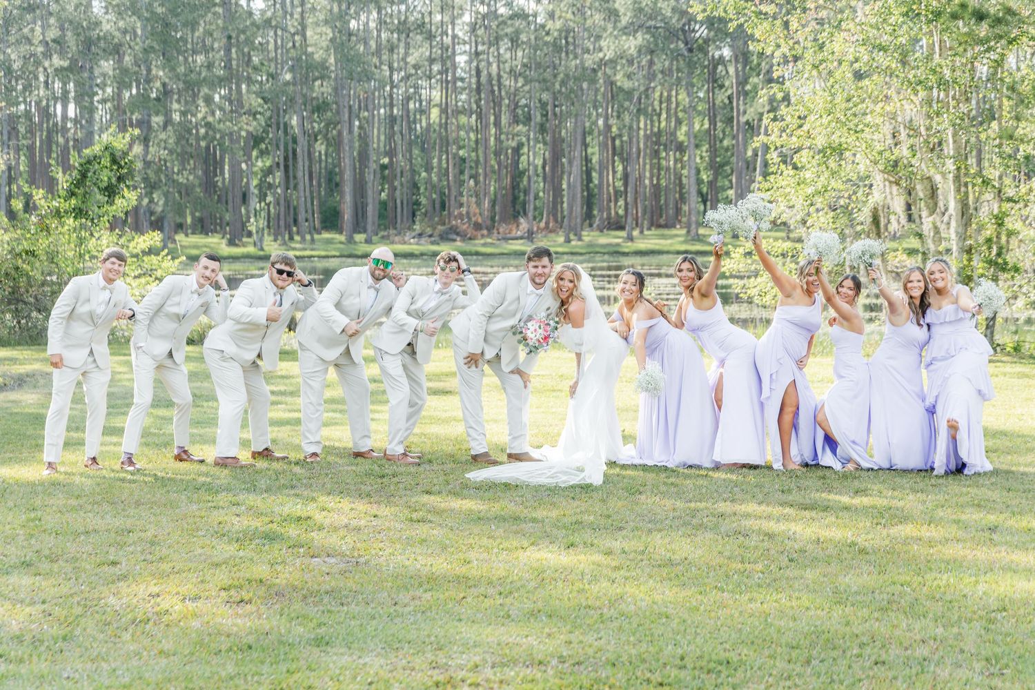 Wedding party forms a line on grass, alternating white suits and lilac dresses in outdoor setting.