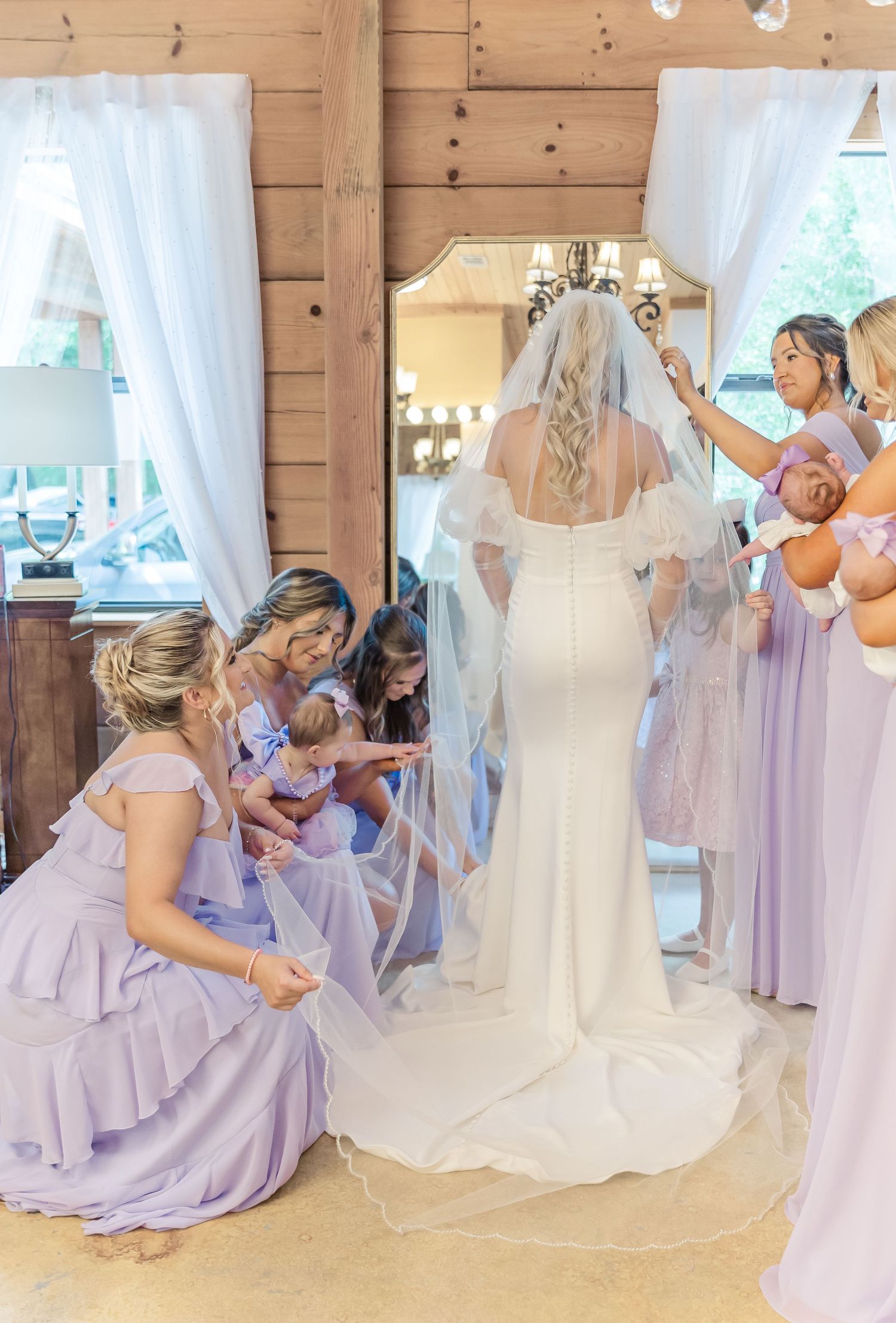 Bridesmaids in purple dresses help arrange the train of a white wedding gown in a wooden room.