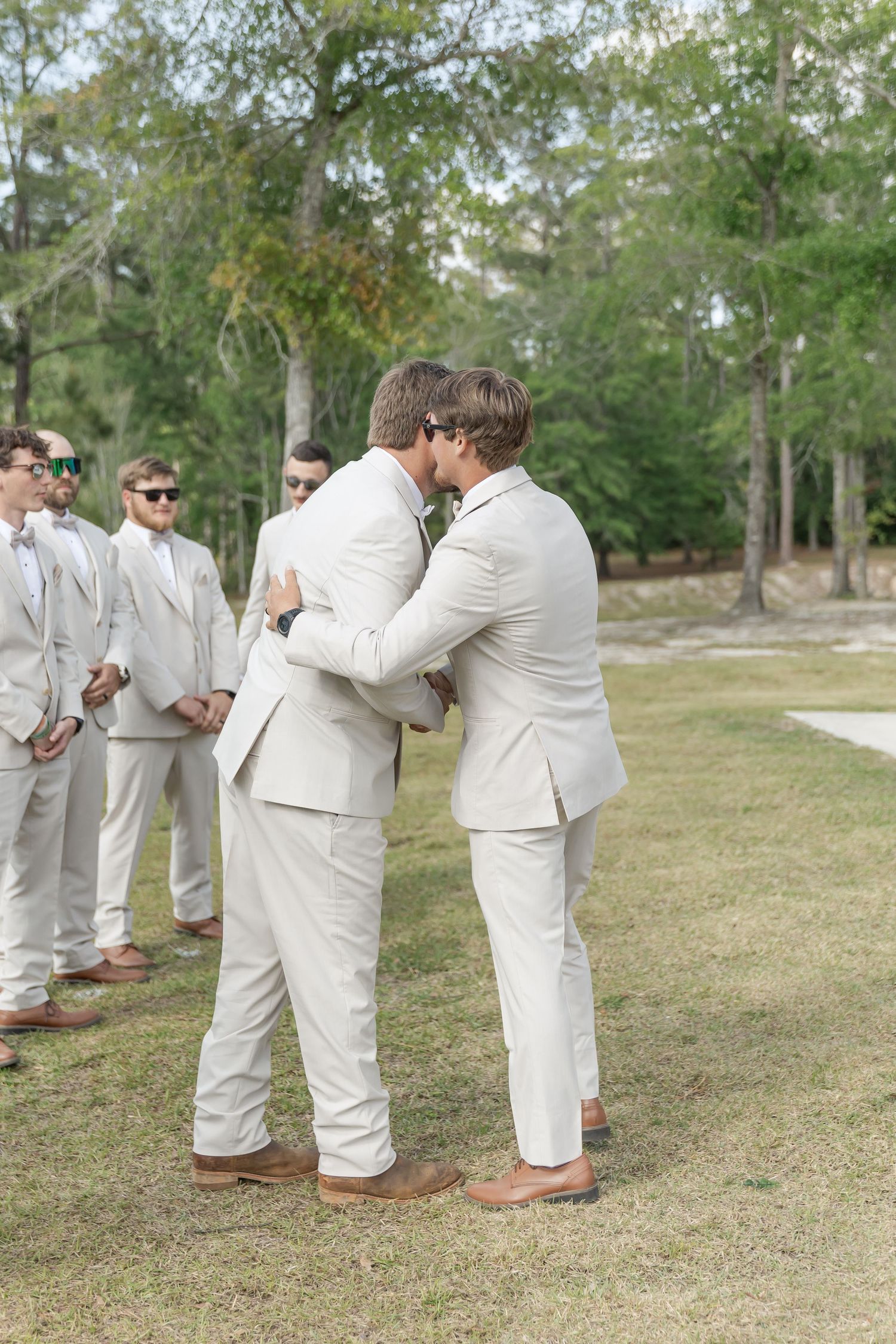Wedding party members in light suits embrace during outdoor ceremony.