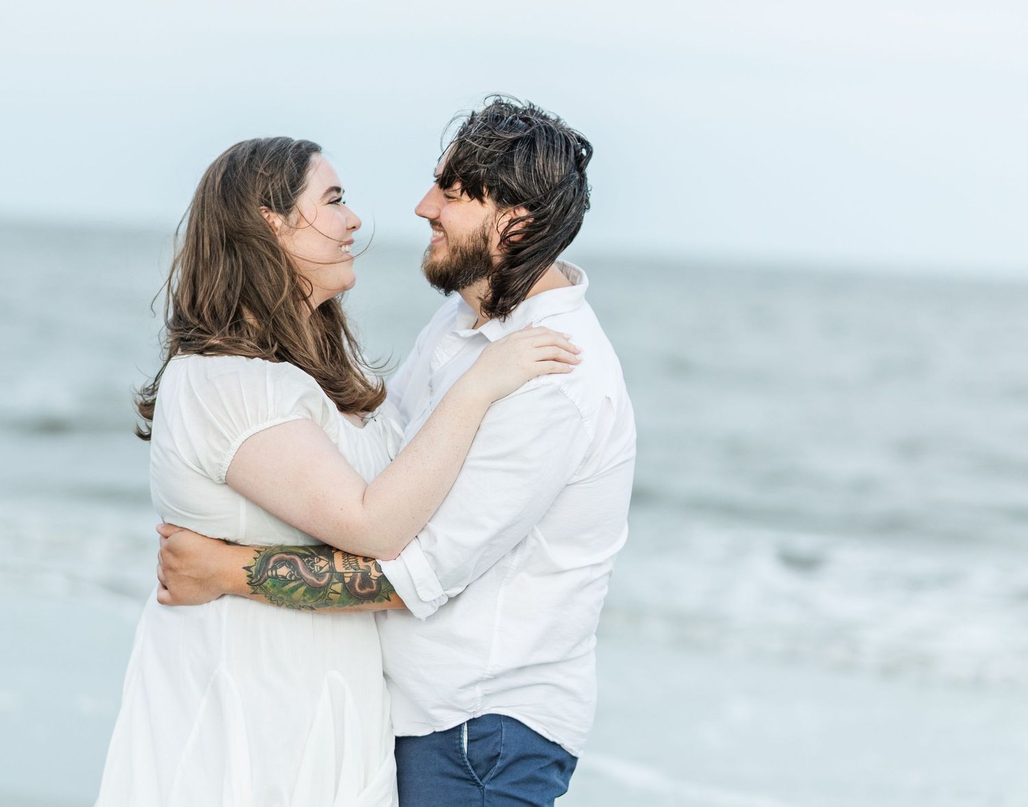 Couple in white embraces on a beach with ocean waves in the background.