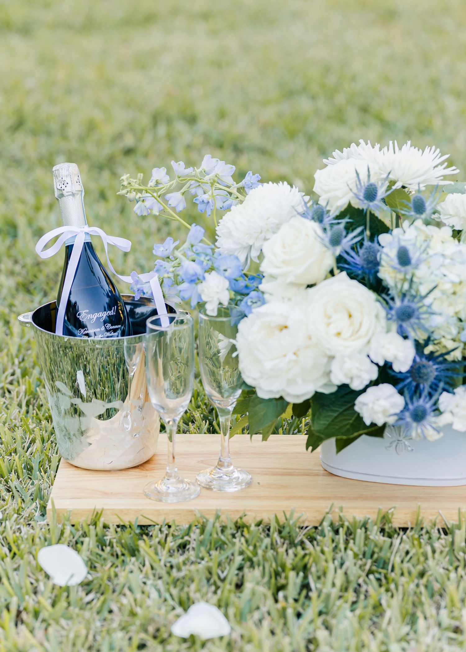 White and blue floral arrangements with roses and glassware displayed on wooden boards in grass.