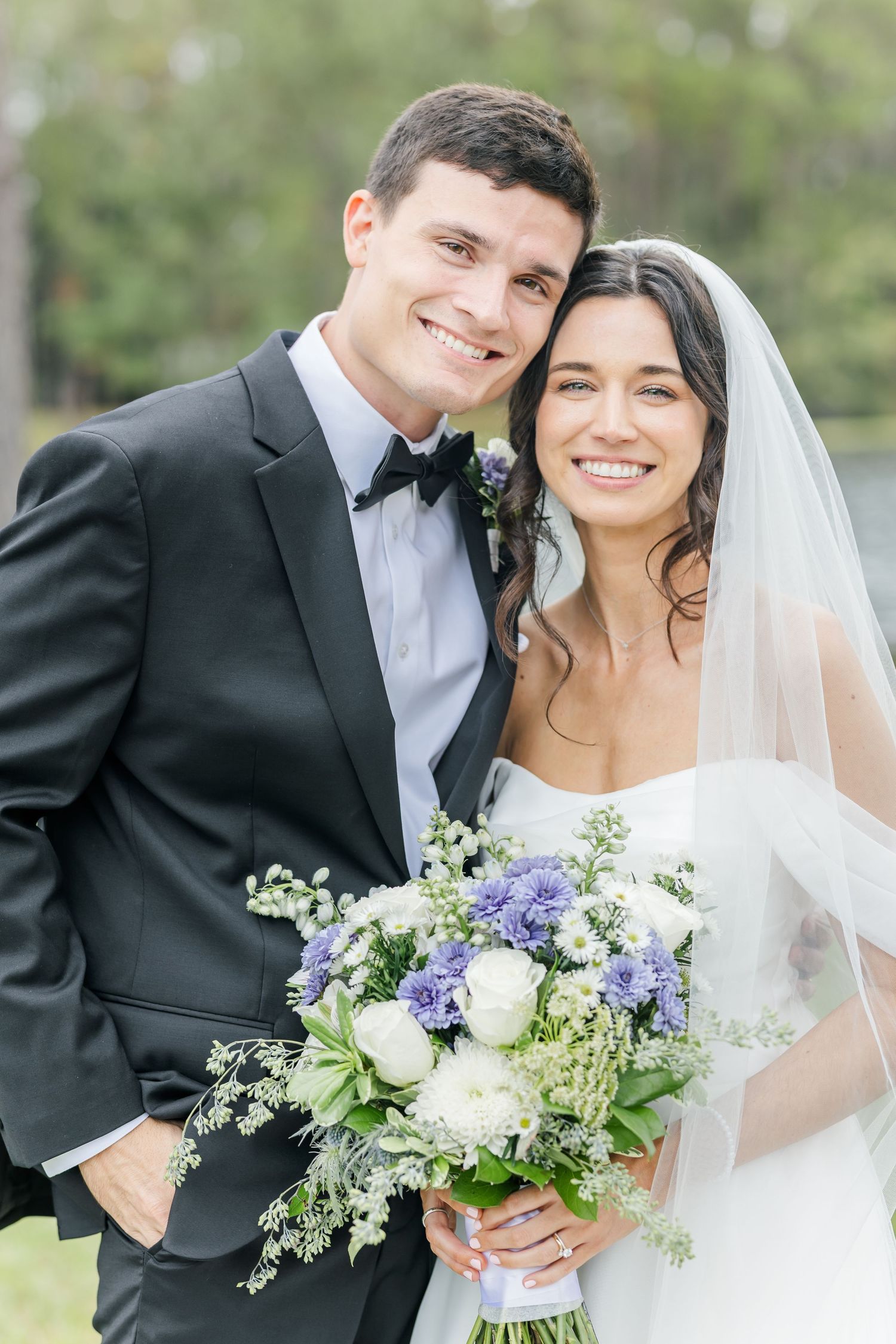 A bride and groom pose together outdoors, with the bride holding a beautiful blue and white floral bouquet.