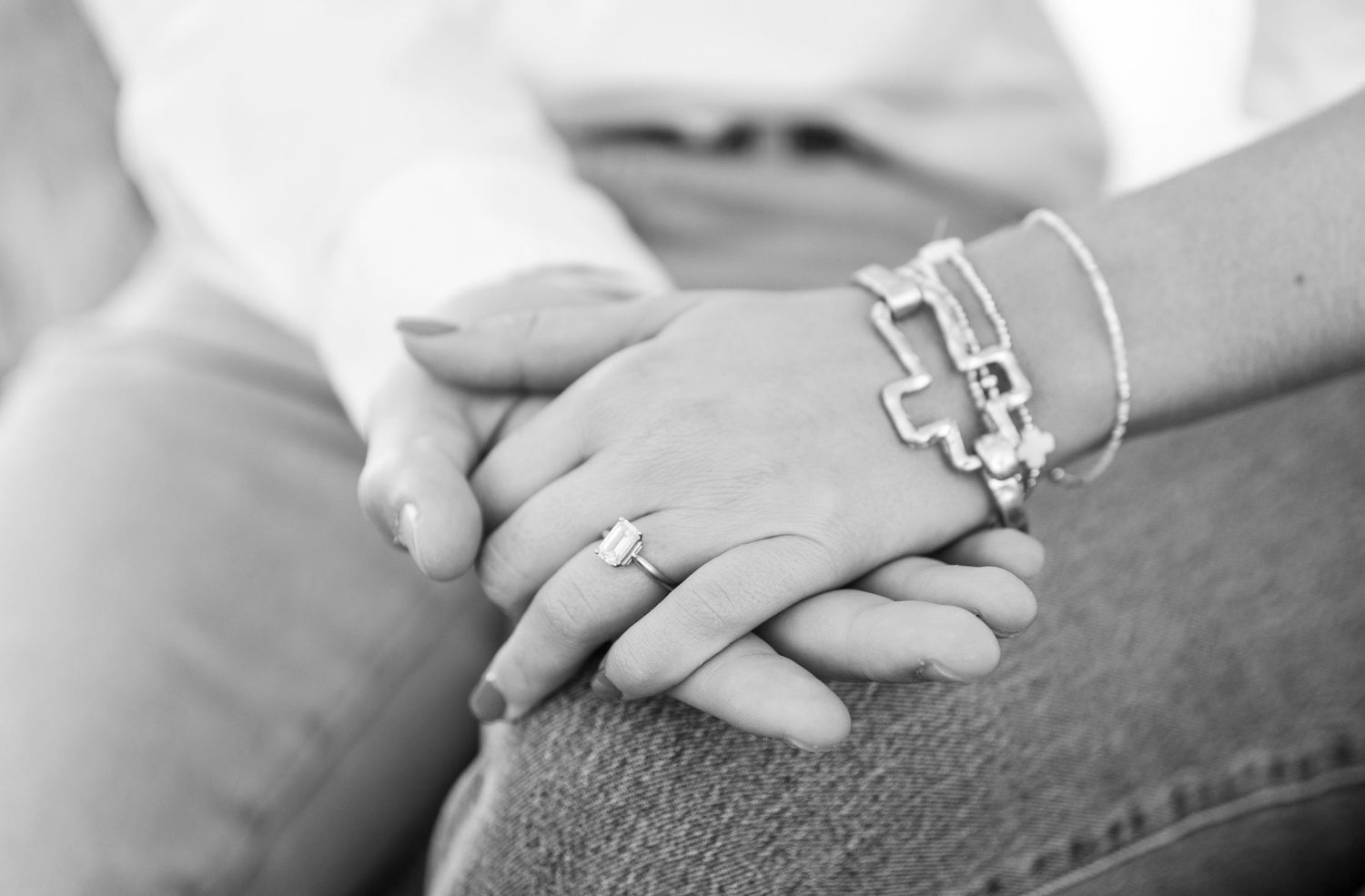 Close-up black and white photo of intertwined hands with engagement ring and bracelets.