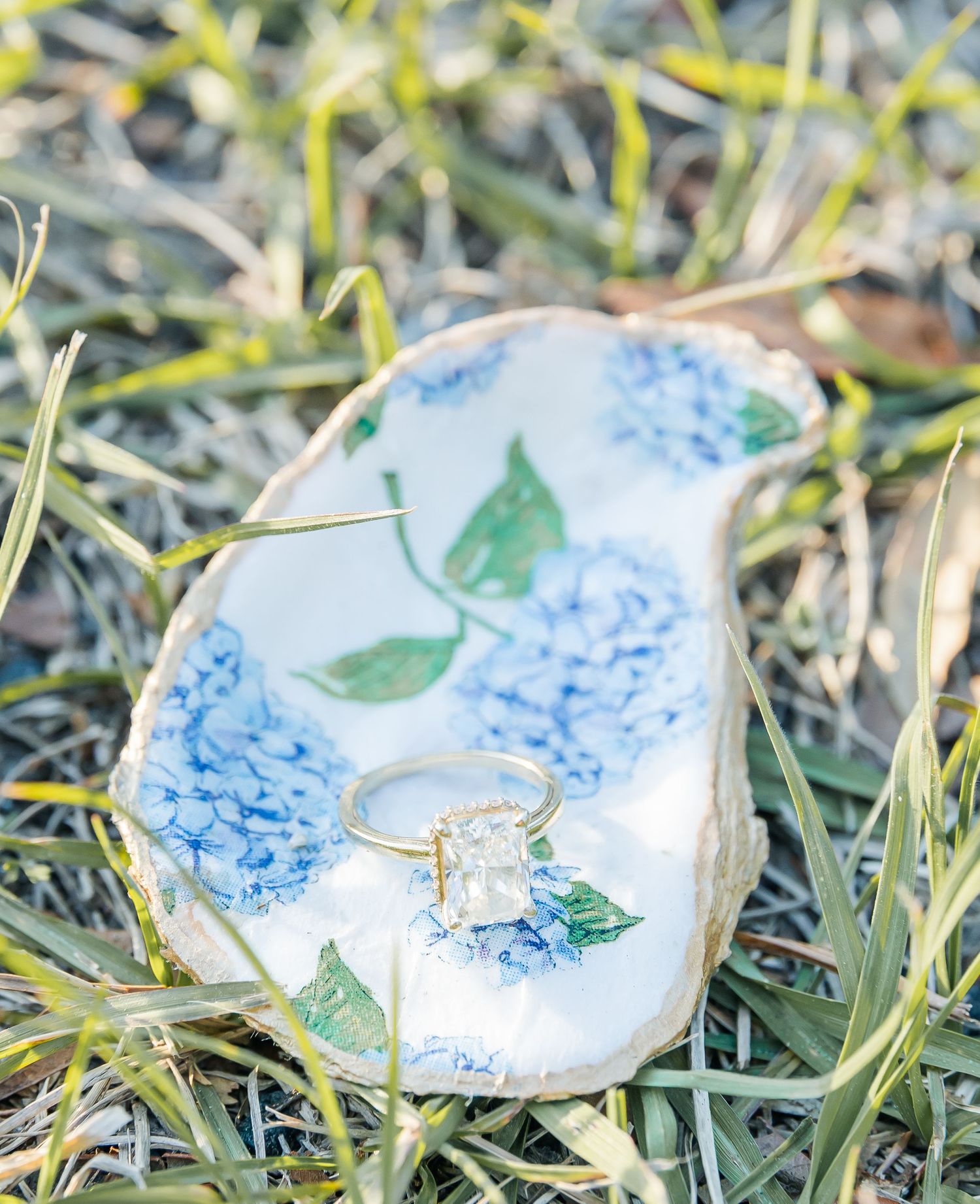 An engagement ring displayed on a decorative dish with blue hydrangea floral pattern laying in grass.
