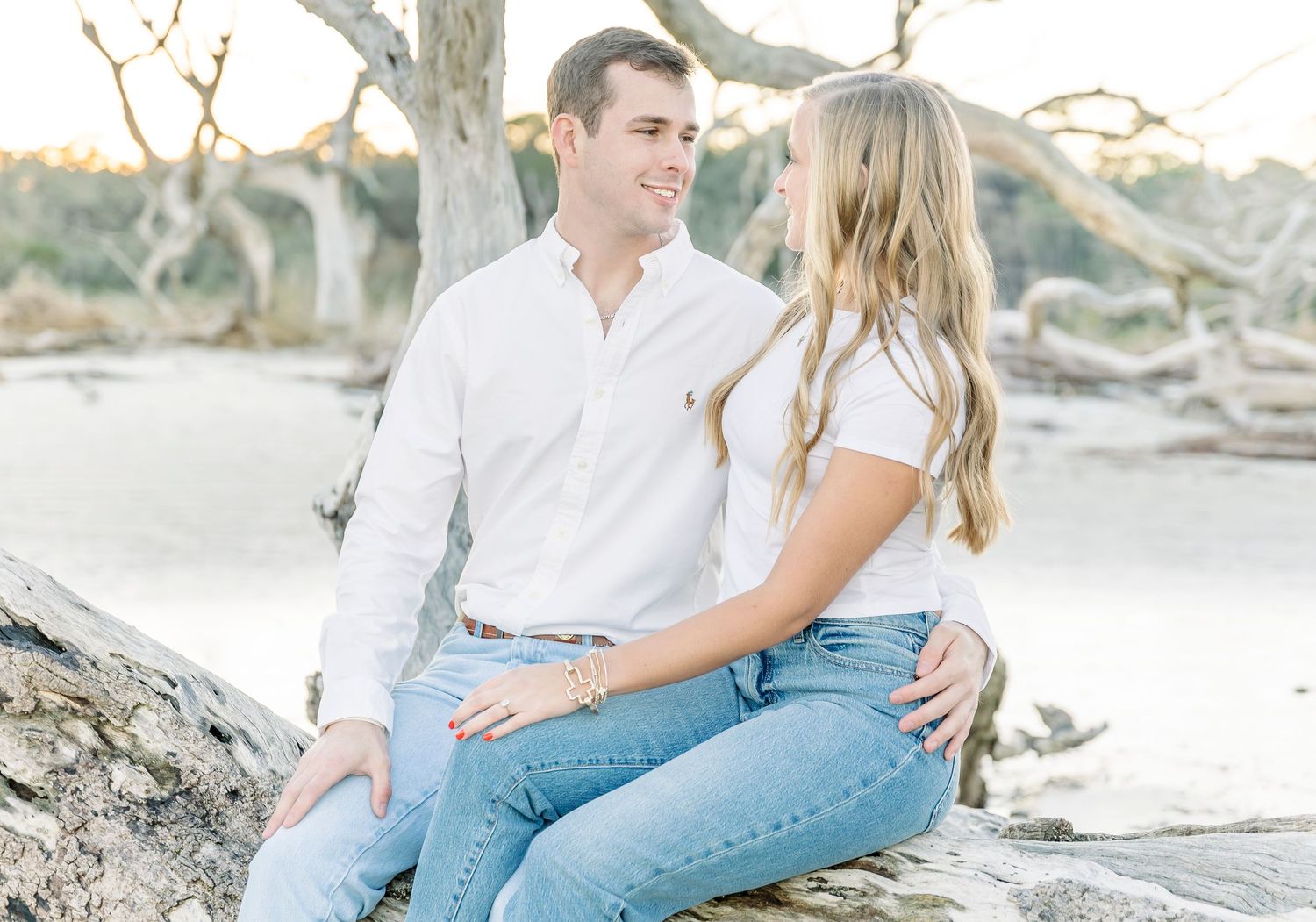 Couple in white tops and jeans sitting together on beach driftwood during sunset.