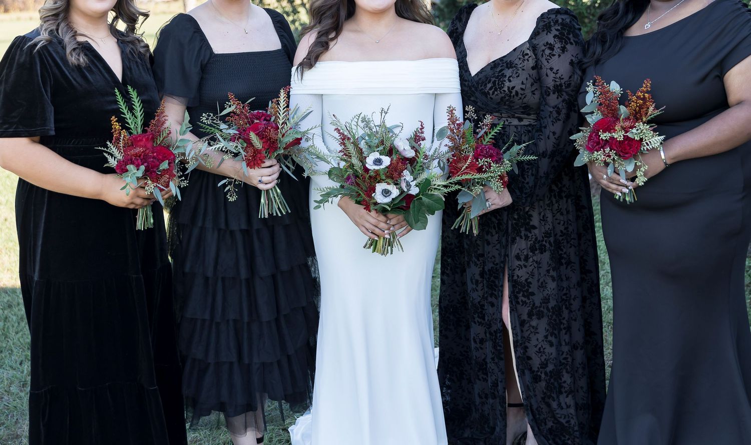 Wedding party members display their red and white bouquets against black dresses outdoors.