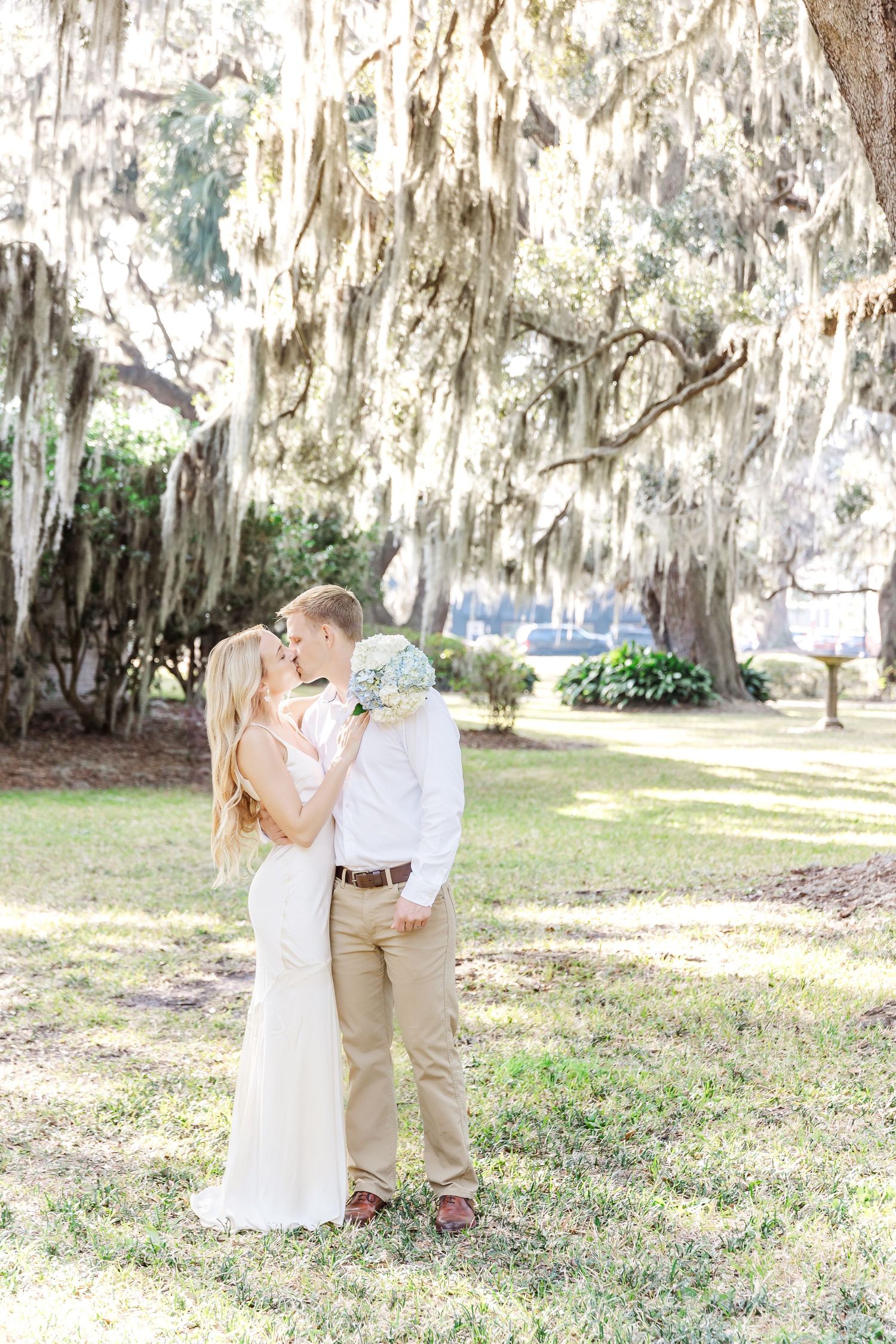 Couple in wedding attire embraces under rustic Spanish moss trees in soft natural lighting.