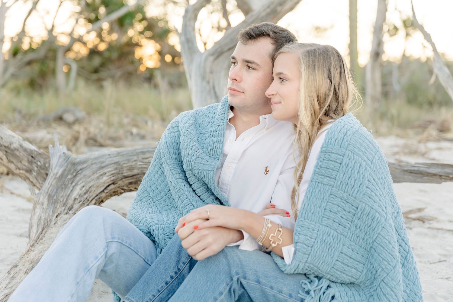 Cozy couple wrapped in blue sweater sitting against driftwood at sunset.