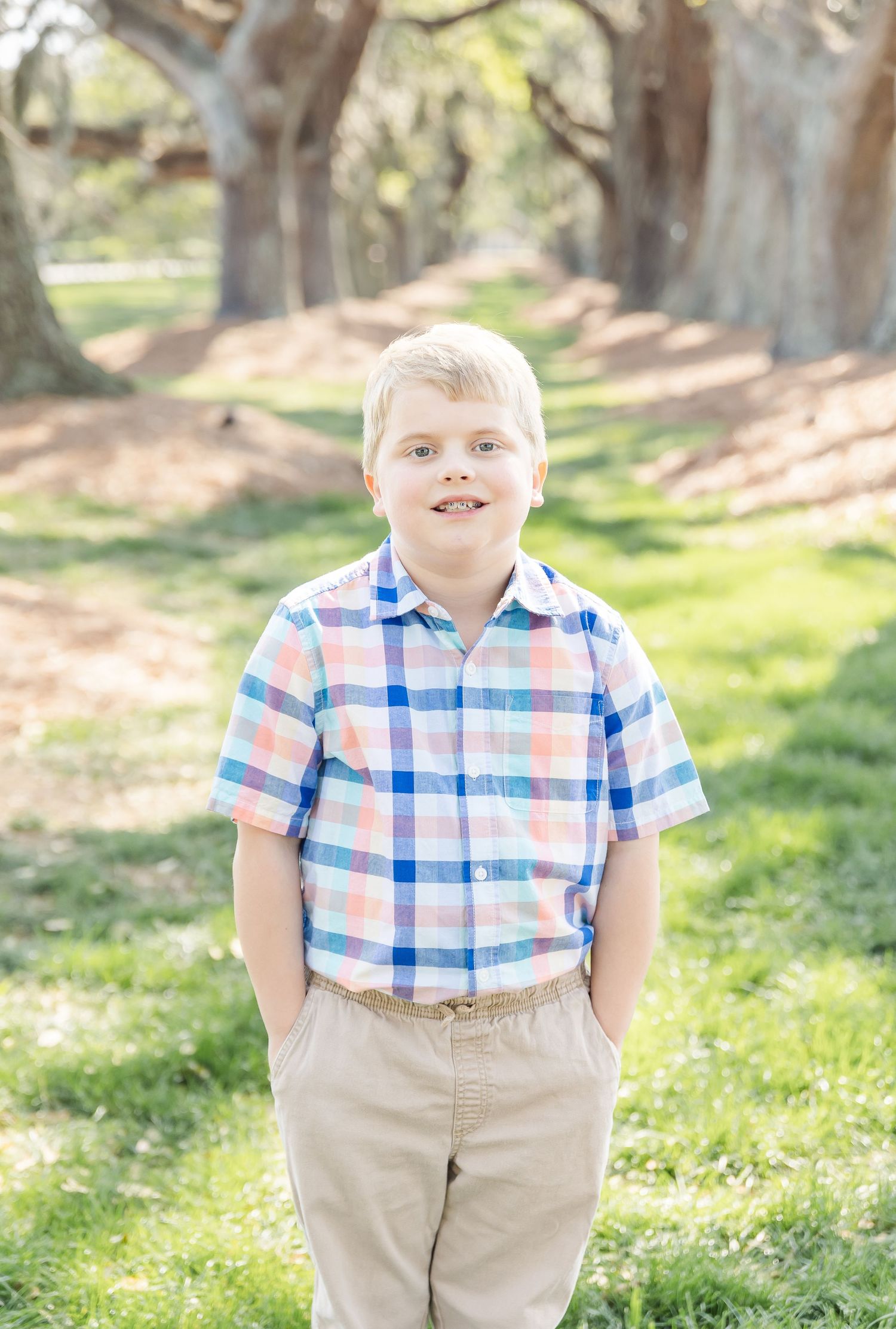 Young boy in plaid shirt and khaki pants smiles at camera while standing under oak trees.