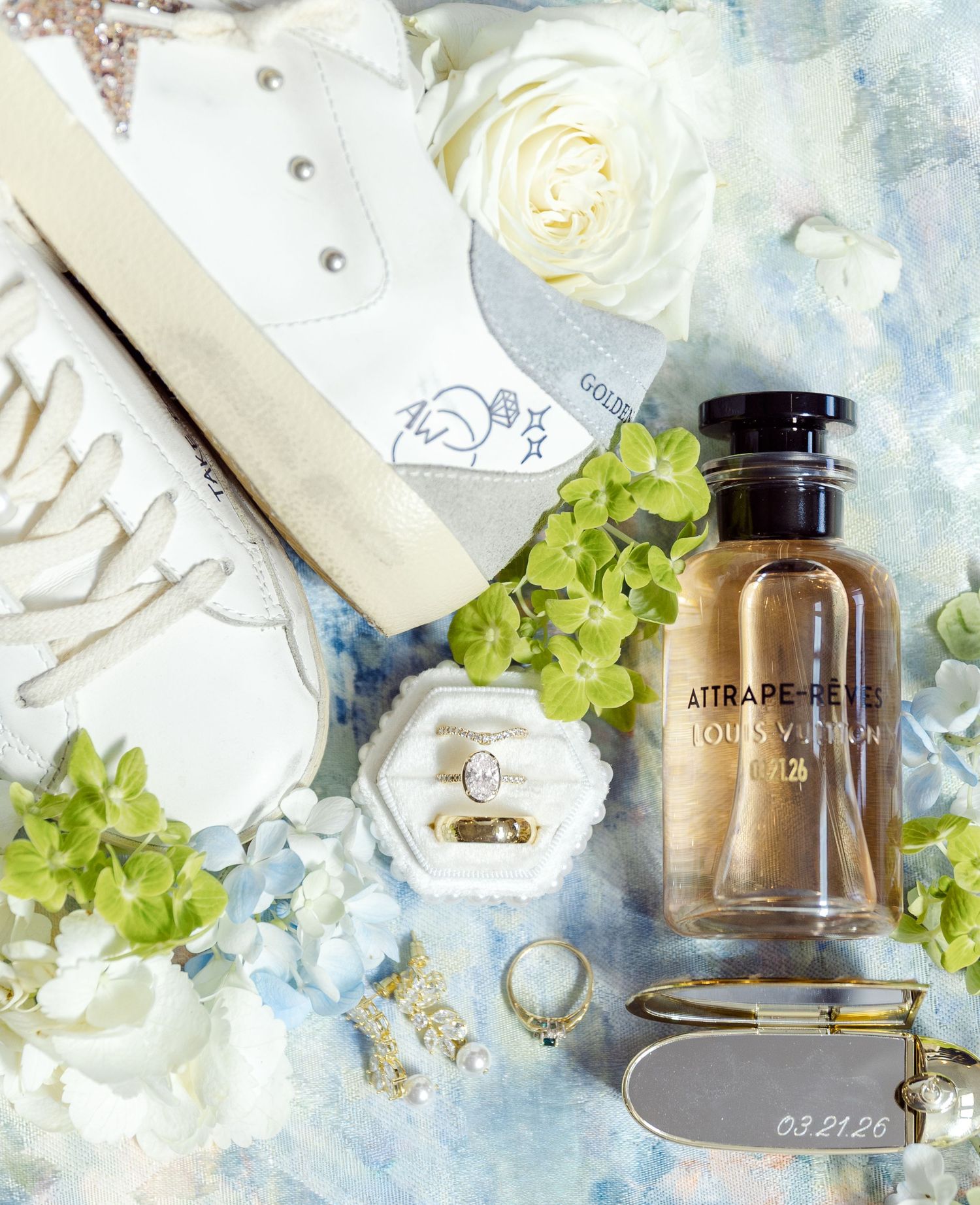 Elegant bridal flat lay with Louis Vuitton perfume, gold rings, white roses, and custom sneakers dated 03.21.25.