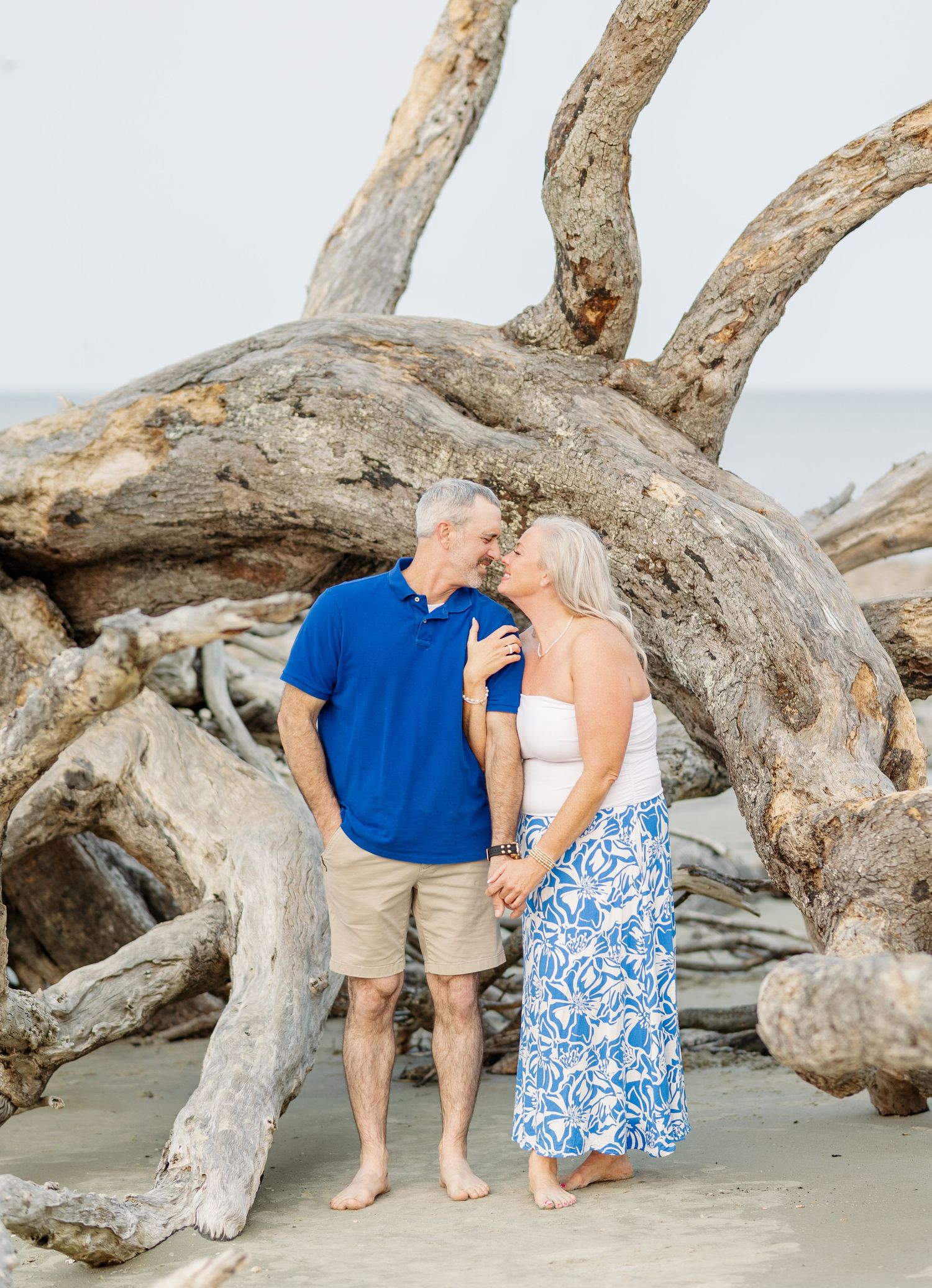 A romantic couple poses along driftwood on a sandy beach in matching blue and white coastal outfits.