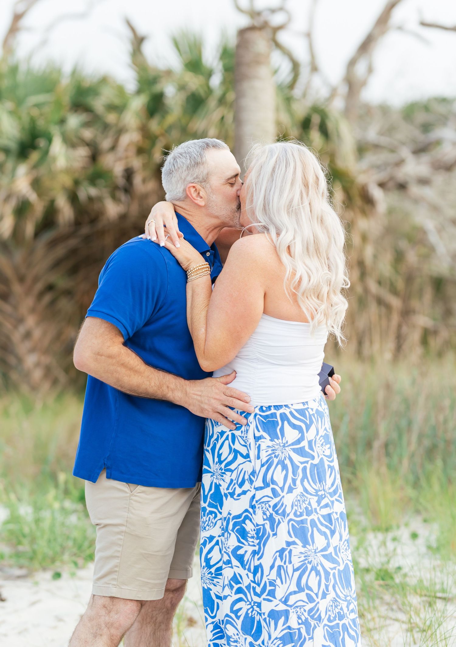 A romantic couple shares a kiss on a sunny beach while dressed in coordinating blue and white outfits.