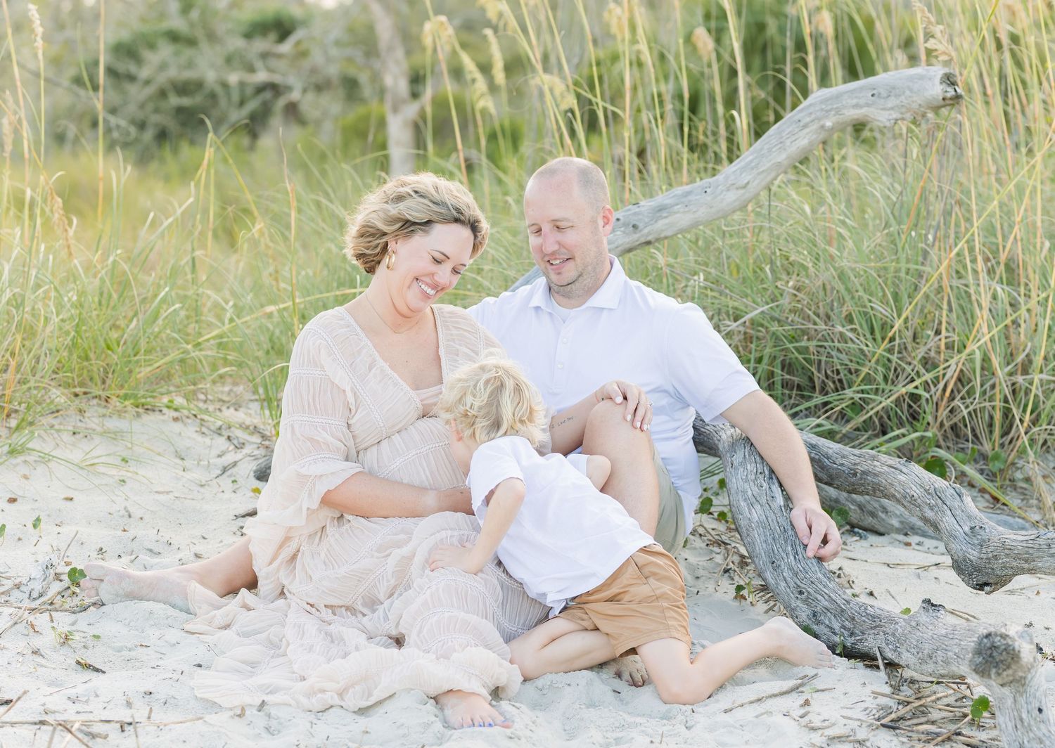 A family relaxes together on a sandy beach surrounded by sea grass and driftwood.