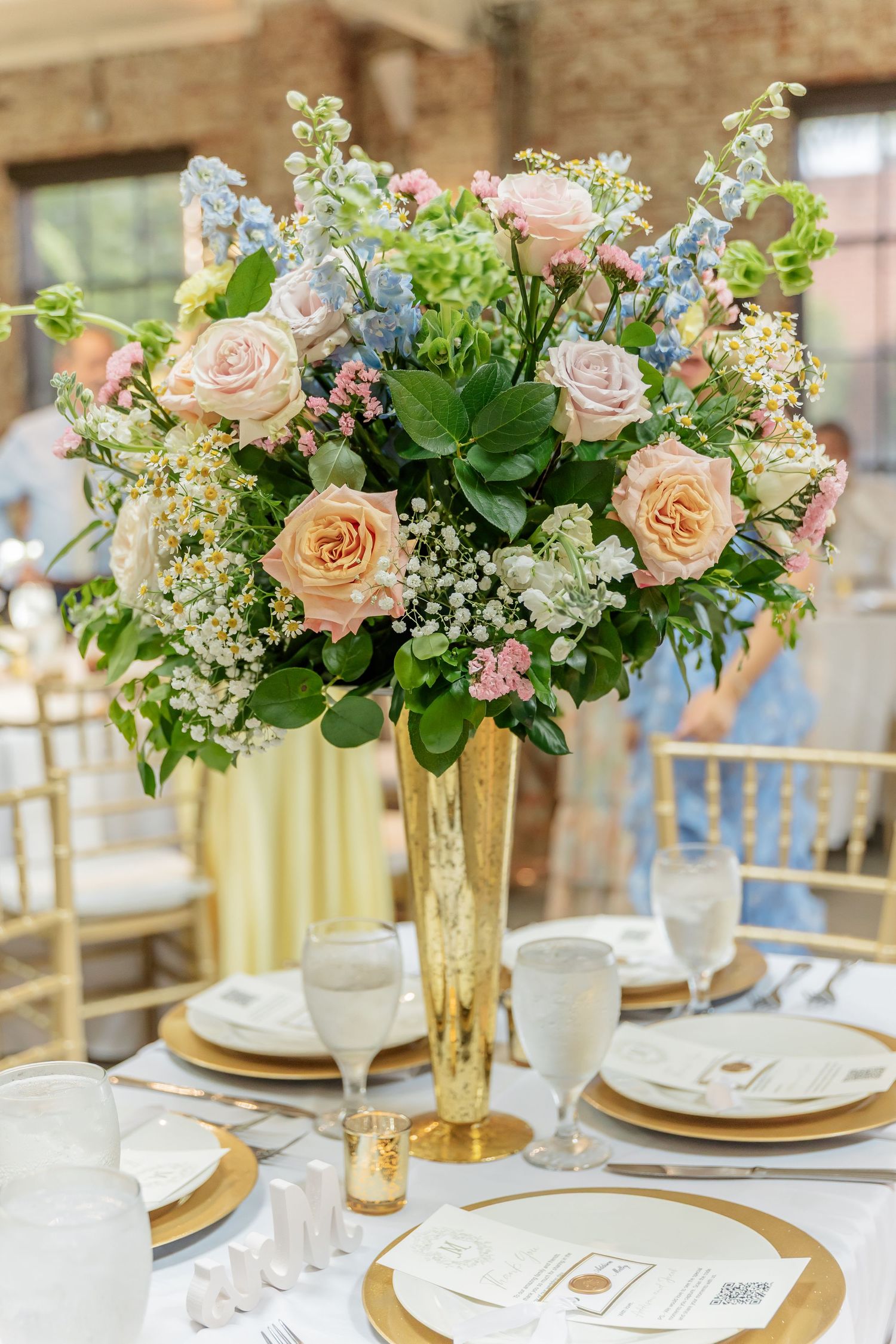Lush floral centerpieces with pink roses and mixed flowers in gold vases on elegant table settings.