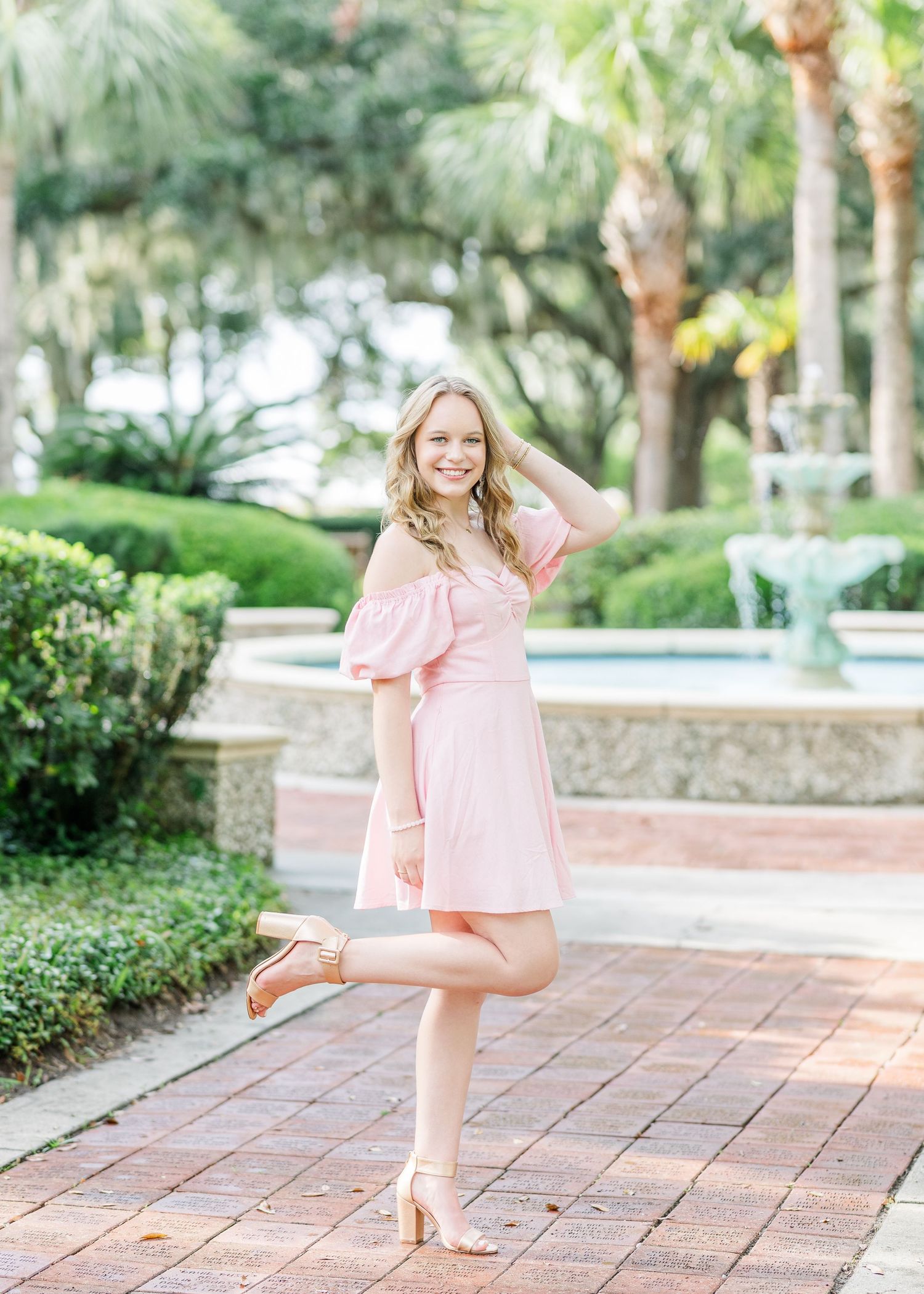 A pink off-shoulder dress stands out against a lush tropical garden and brick pathway on a sunny day.