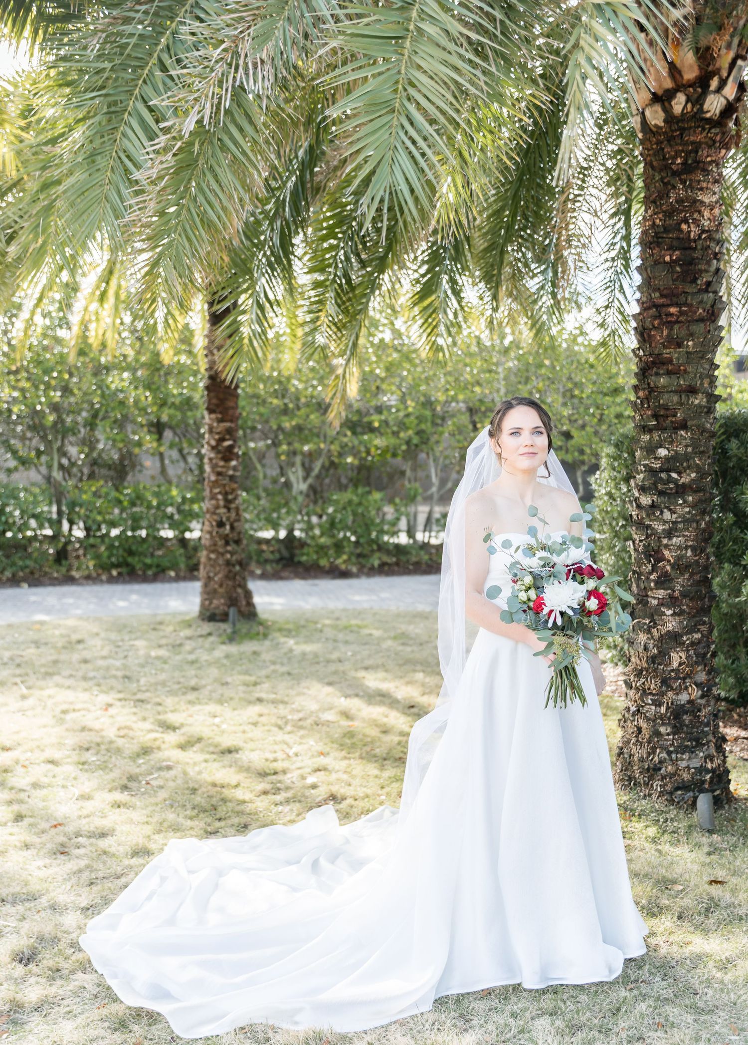 Long white wedding dress train spread elegantly among palm trees outdoors.