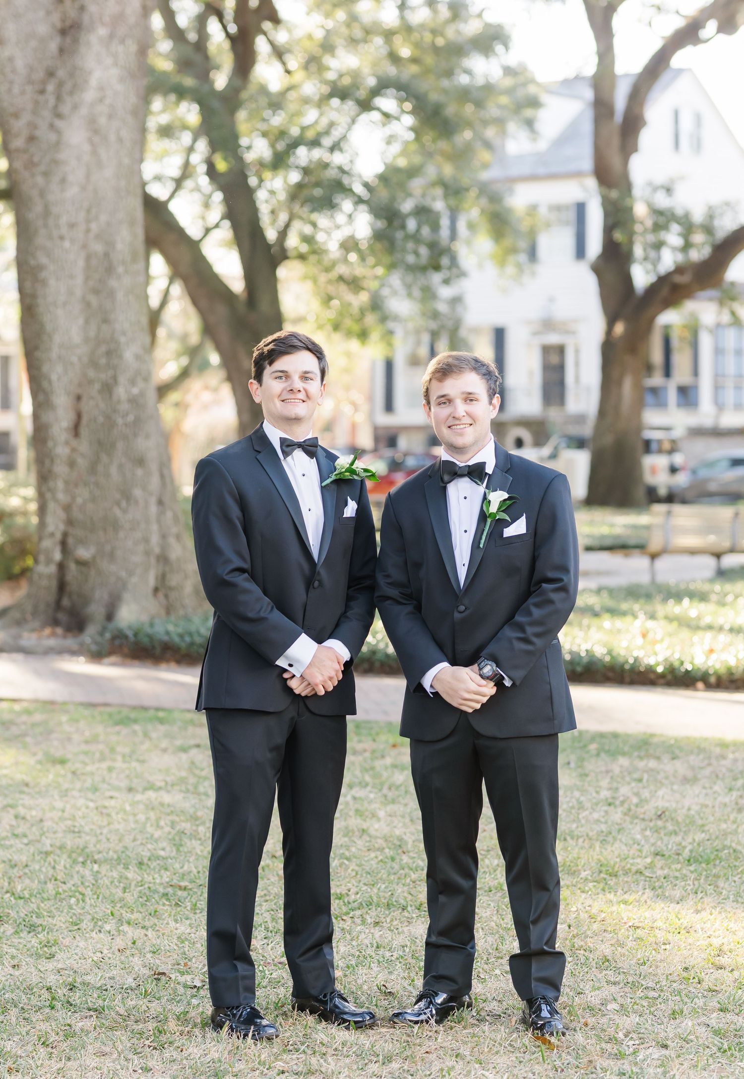 Two groomsmen in classic black tuxedos with bow ties and boutonnieres pose together outdoors under large trees.