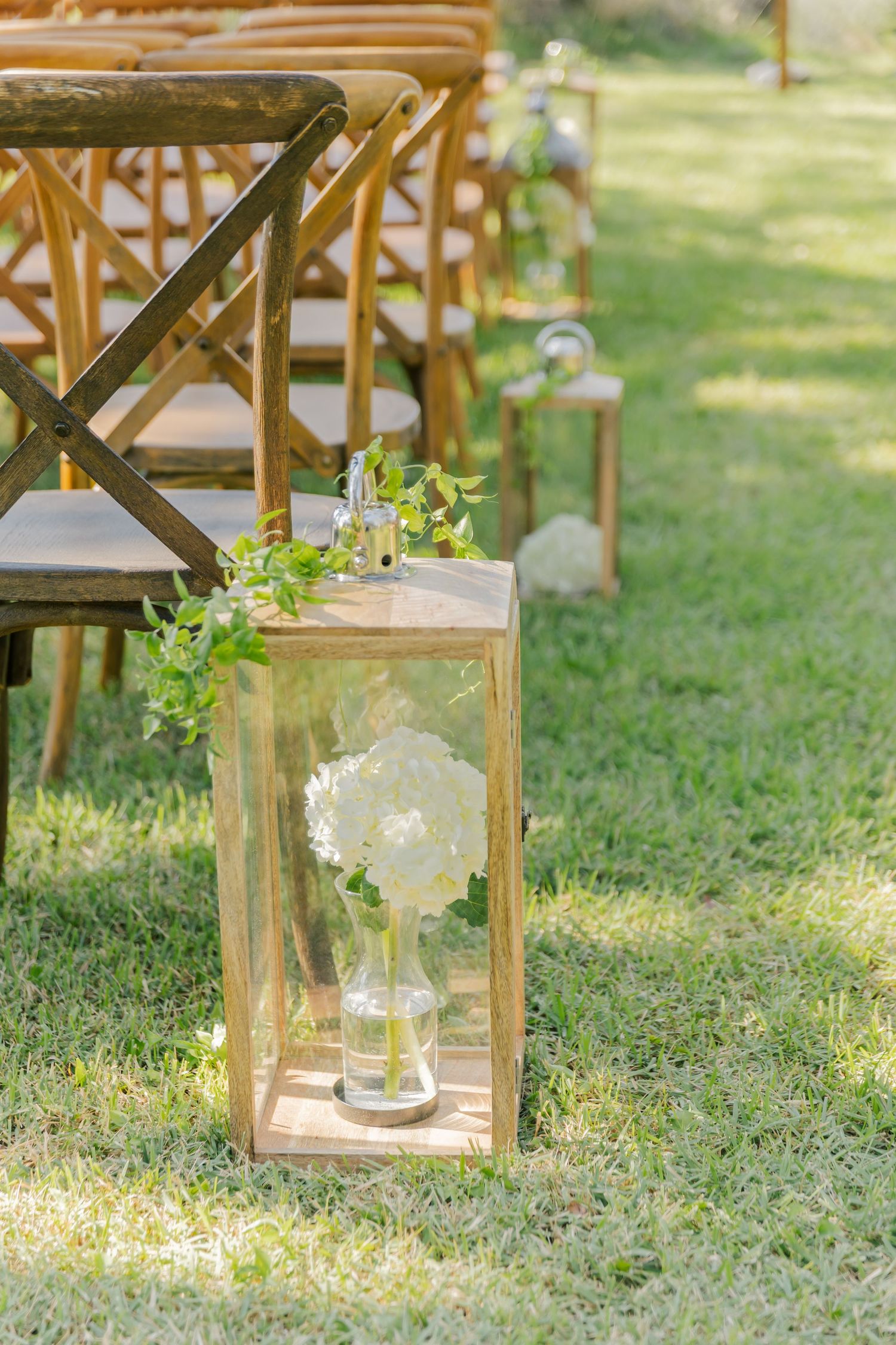 Rustic wooden lanterns with white floral arrangements line a grassy outdoor wedding aisle.