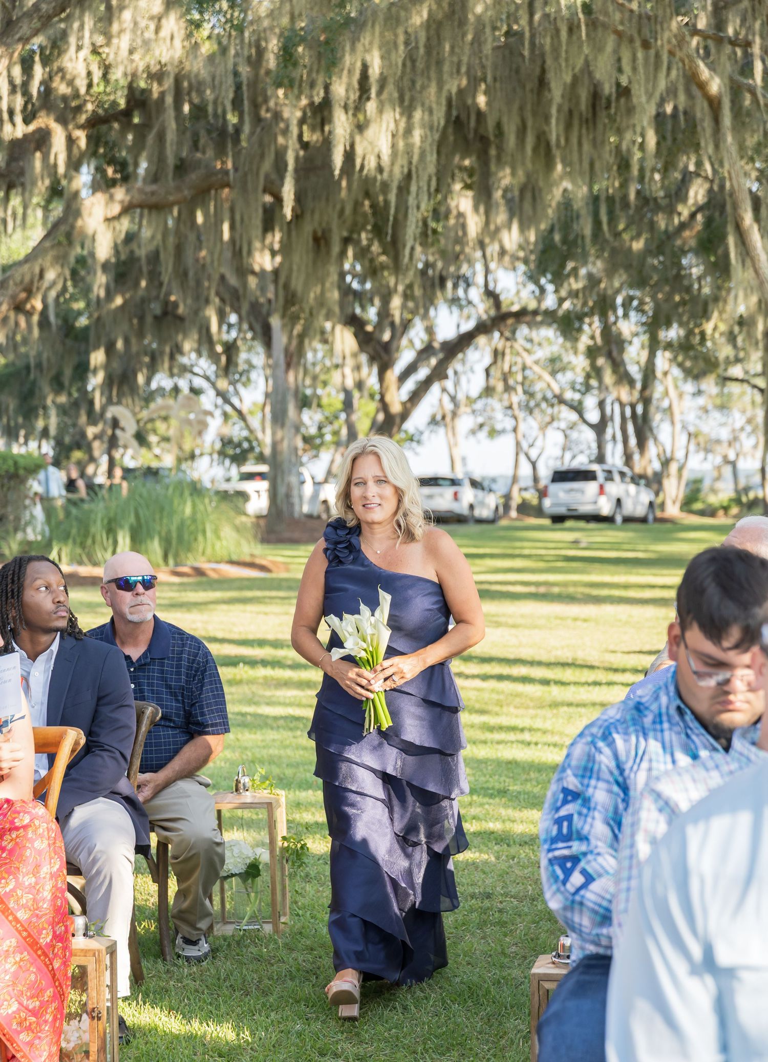 Wedding guests seated on wooden chairs under moss-covered trees during an outdoor ceremony.