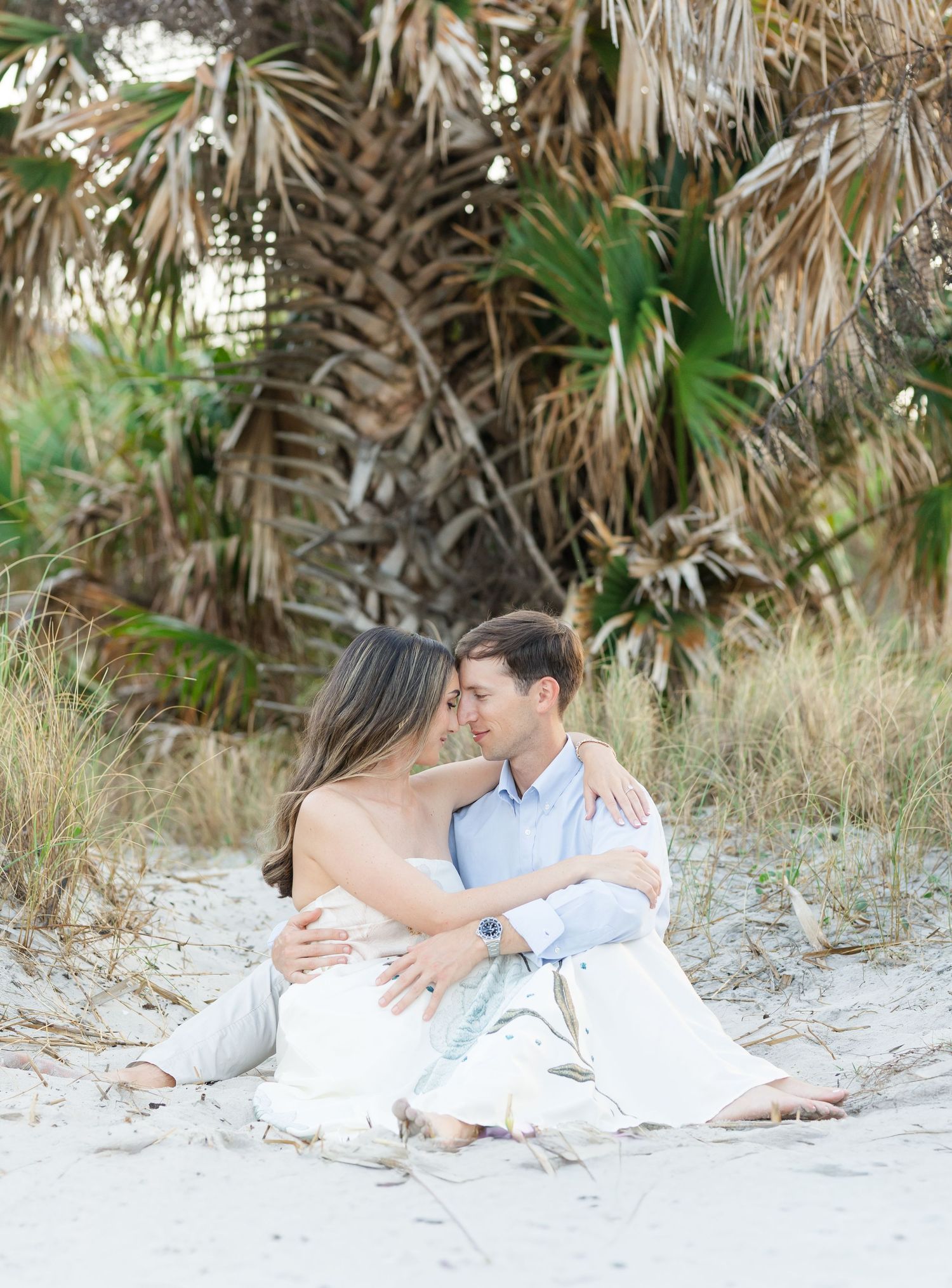 Couple embracing on sandy beach with lush palm tree backdrop, sharing a tender romantic moment.