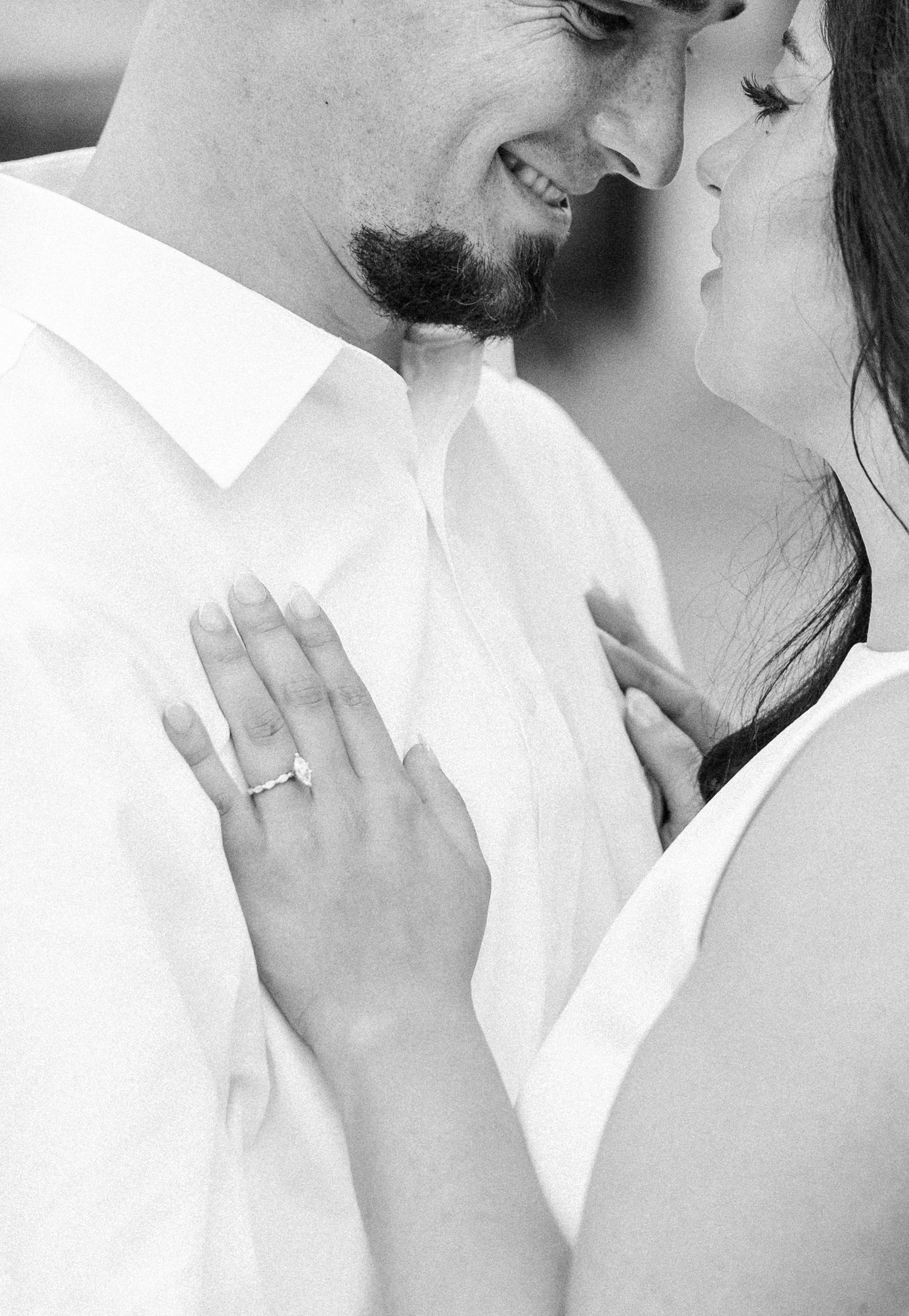 Black and white close-up of smiling couple nearly kissing, woman showing diamond engagement ring.