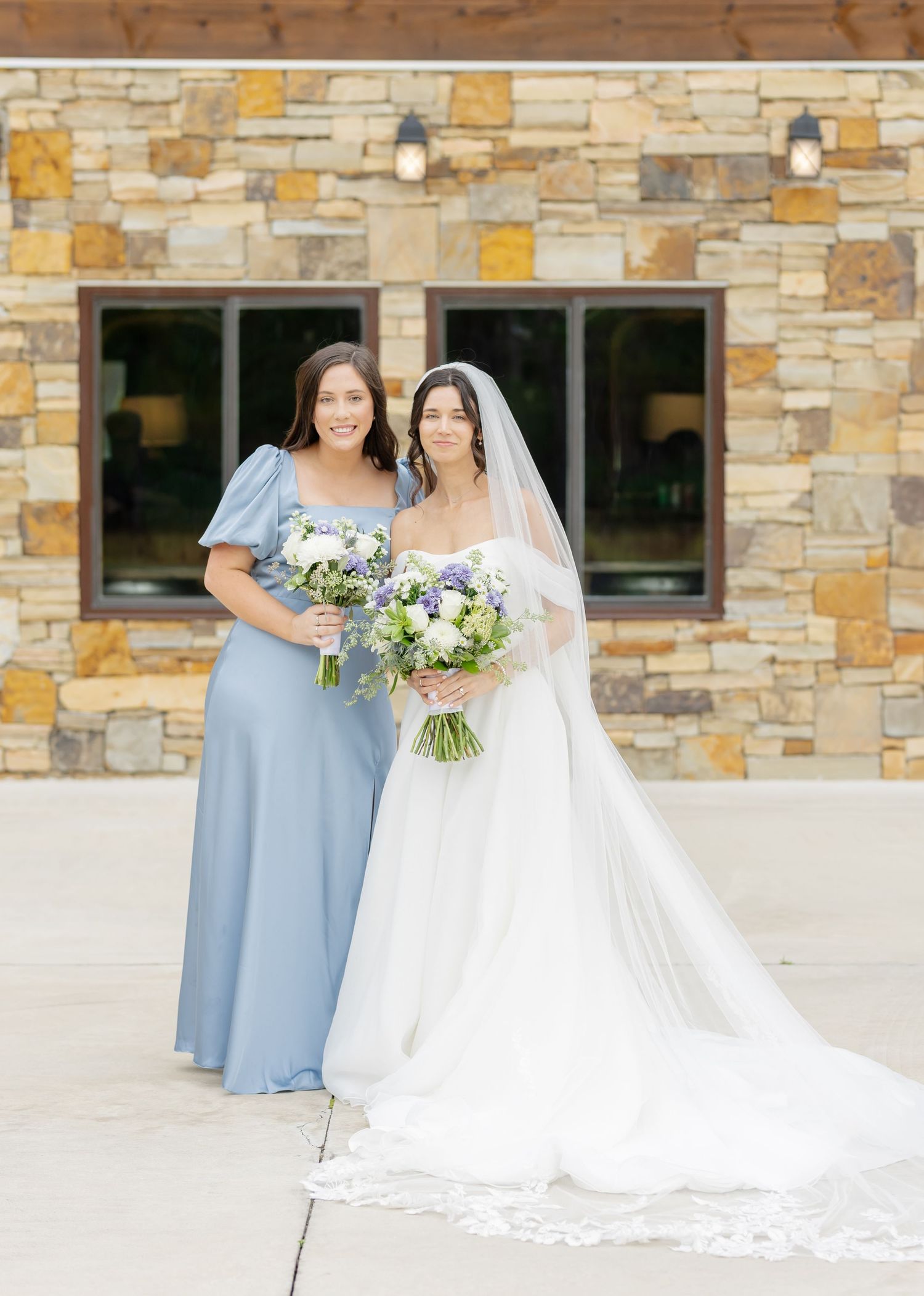 Bridesmaids in powder blue dresses stand with bride in flowing white gown against stone building.