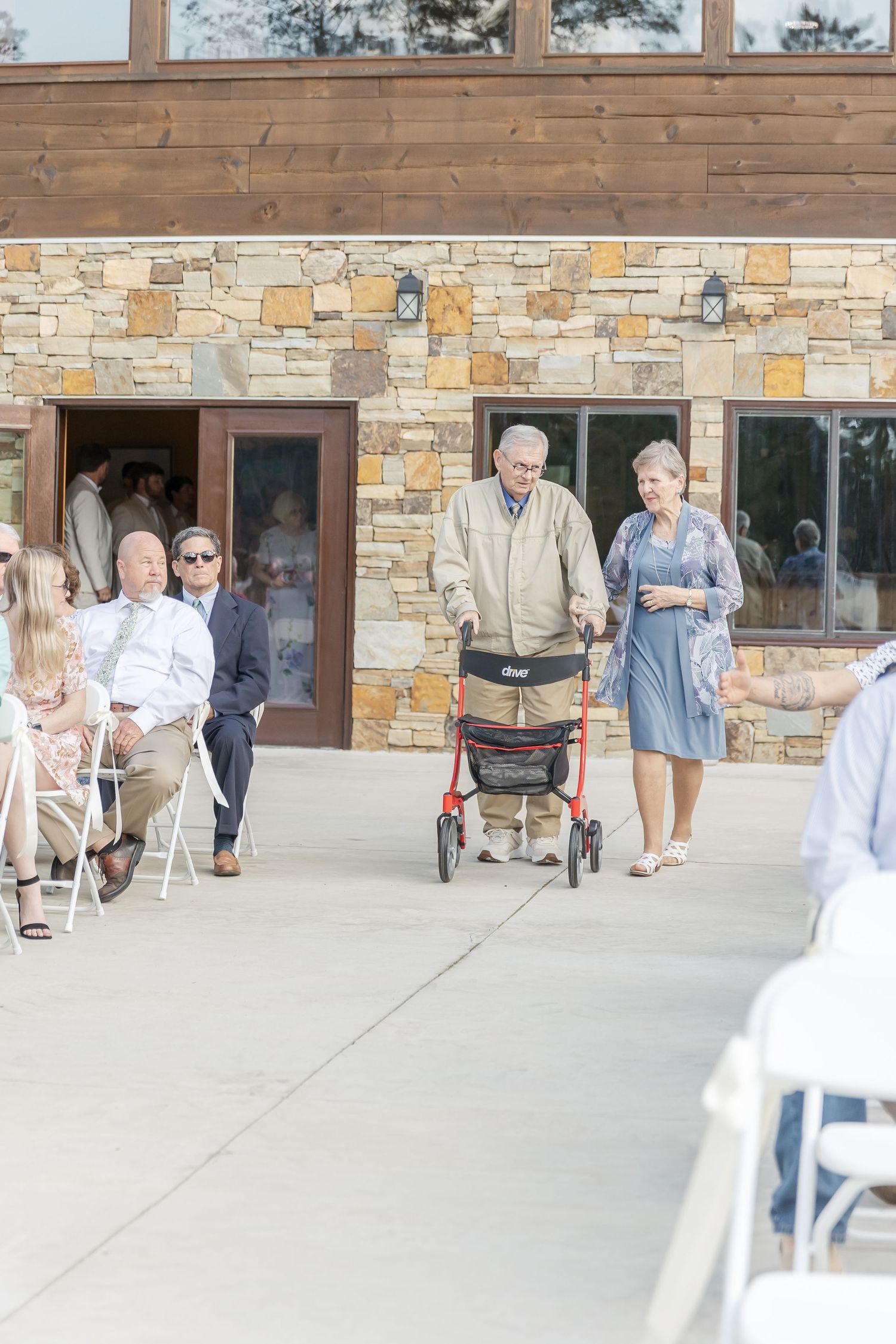 Outdoor processional at stone and wood venue with guests seated in white chairs.