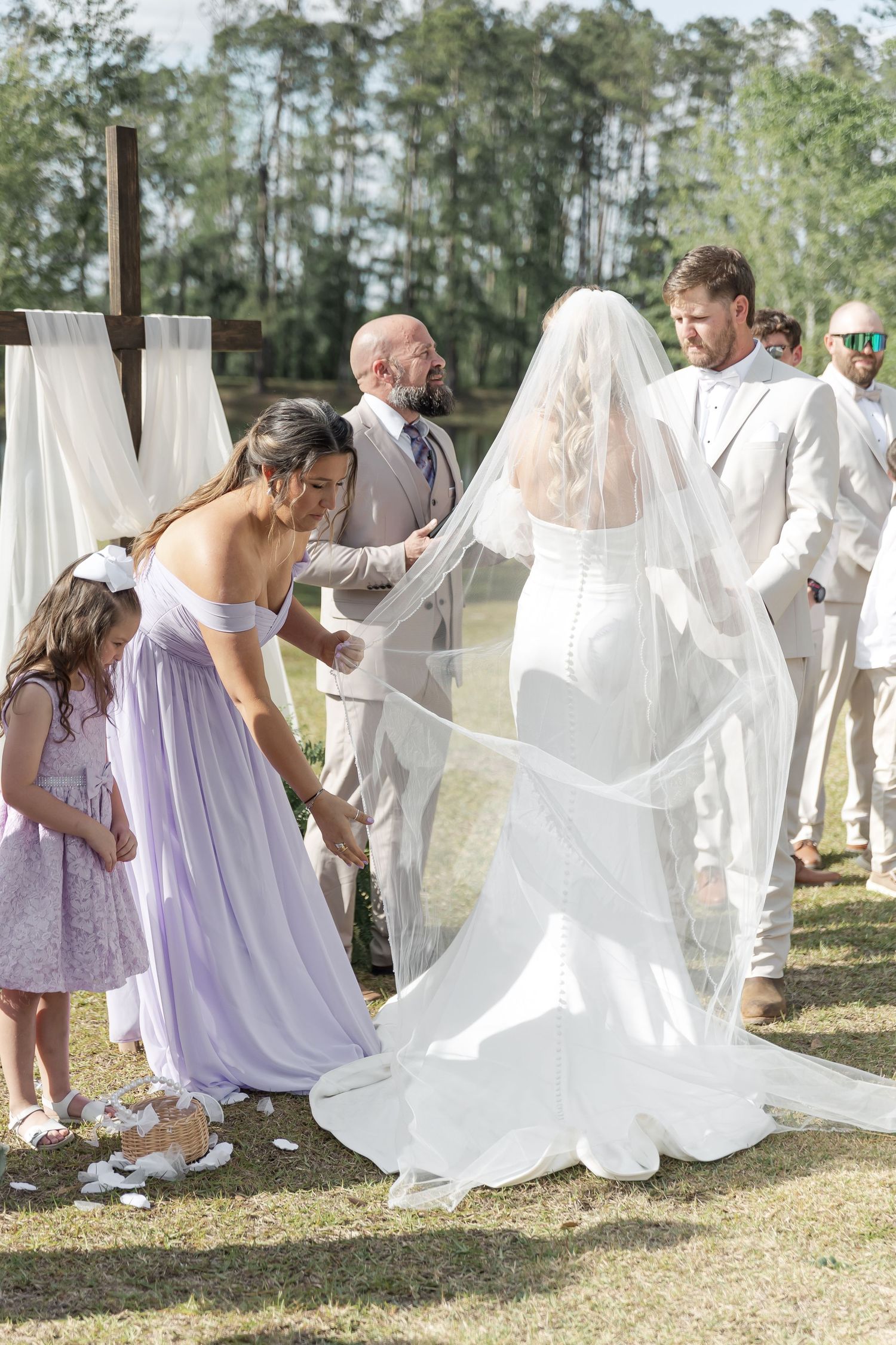 Wedding party helps adjust bride's train during outdoor ceremony by pine trees.