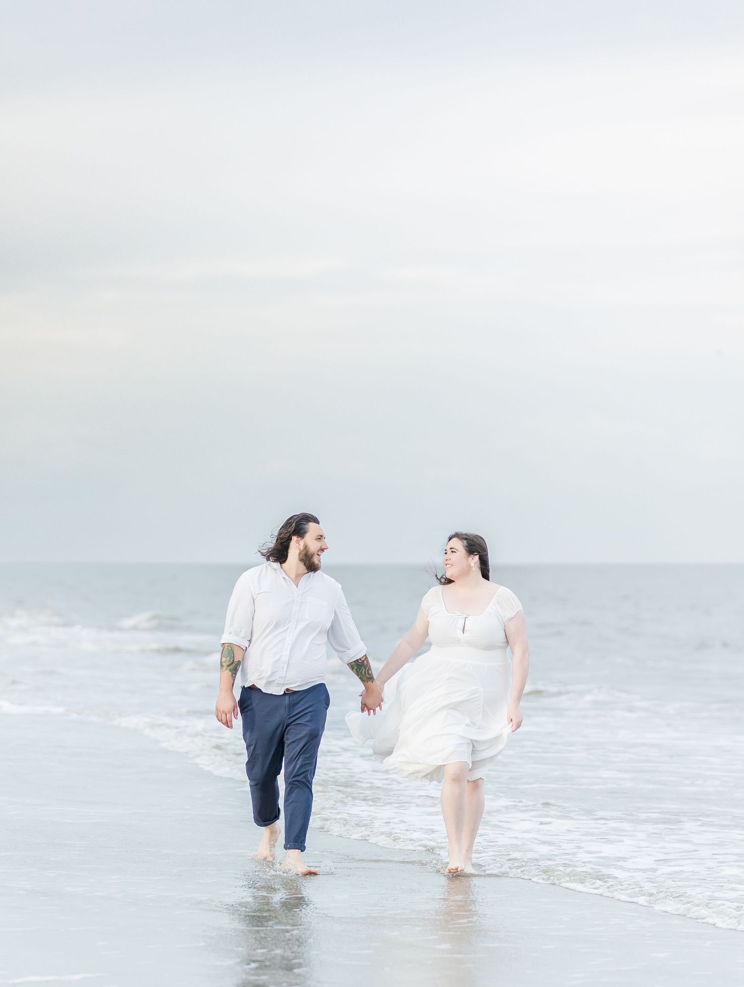 Couple holding hands while walking through gentle waves on a misty beach.