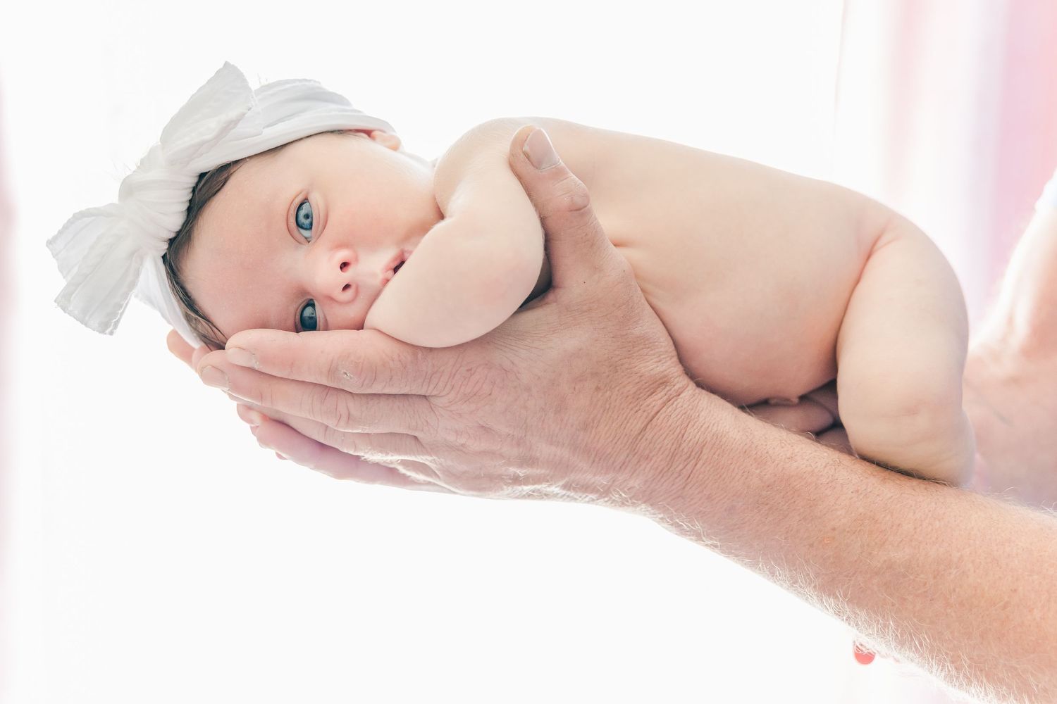 Newborn baby with blue eyes wearing white bow held gently in hands.