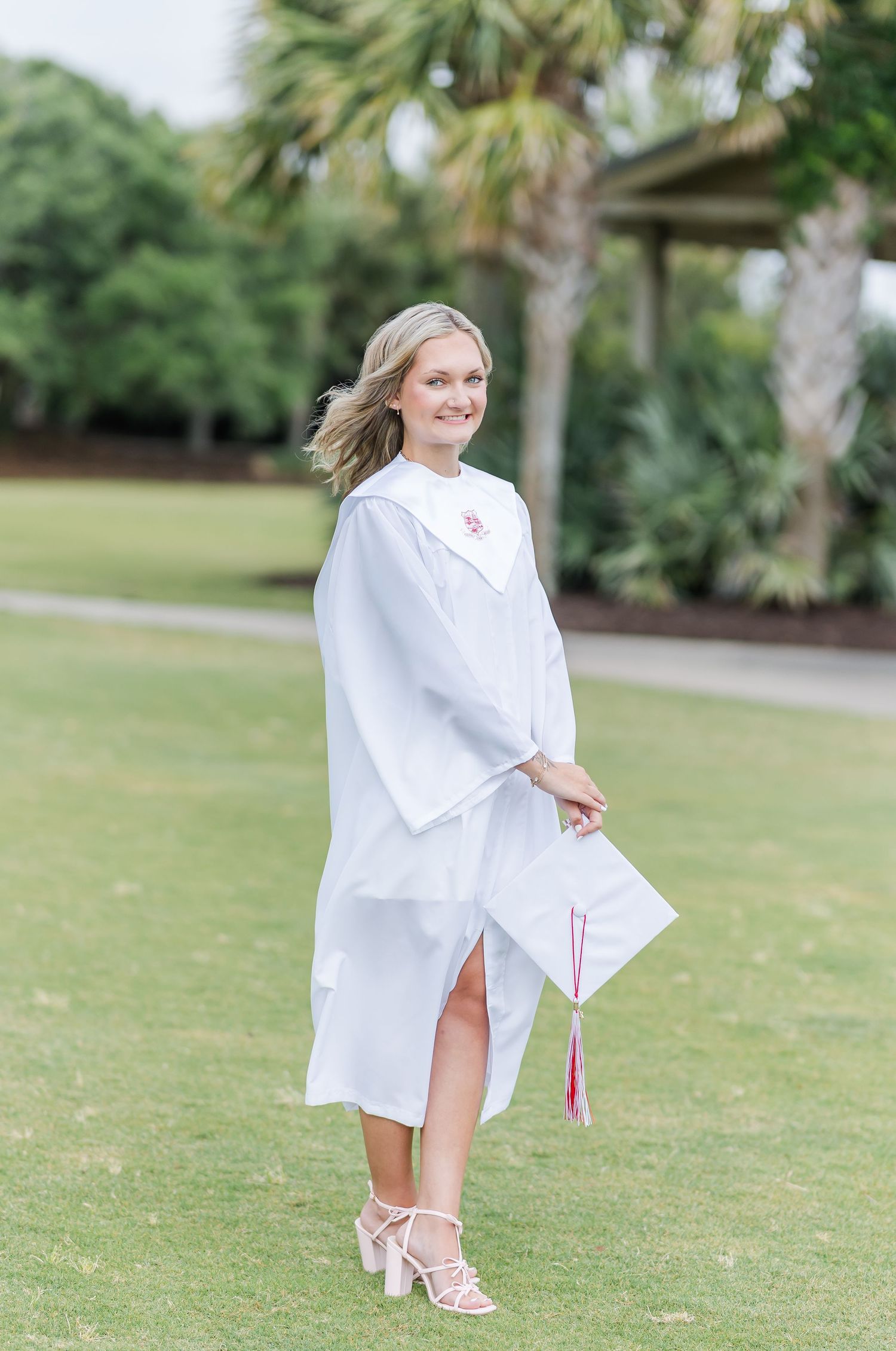 A person in a white graduation gown poses outdoors near palm trees on a grassy lawn.