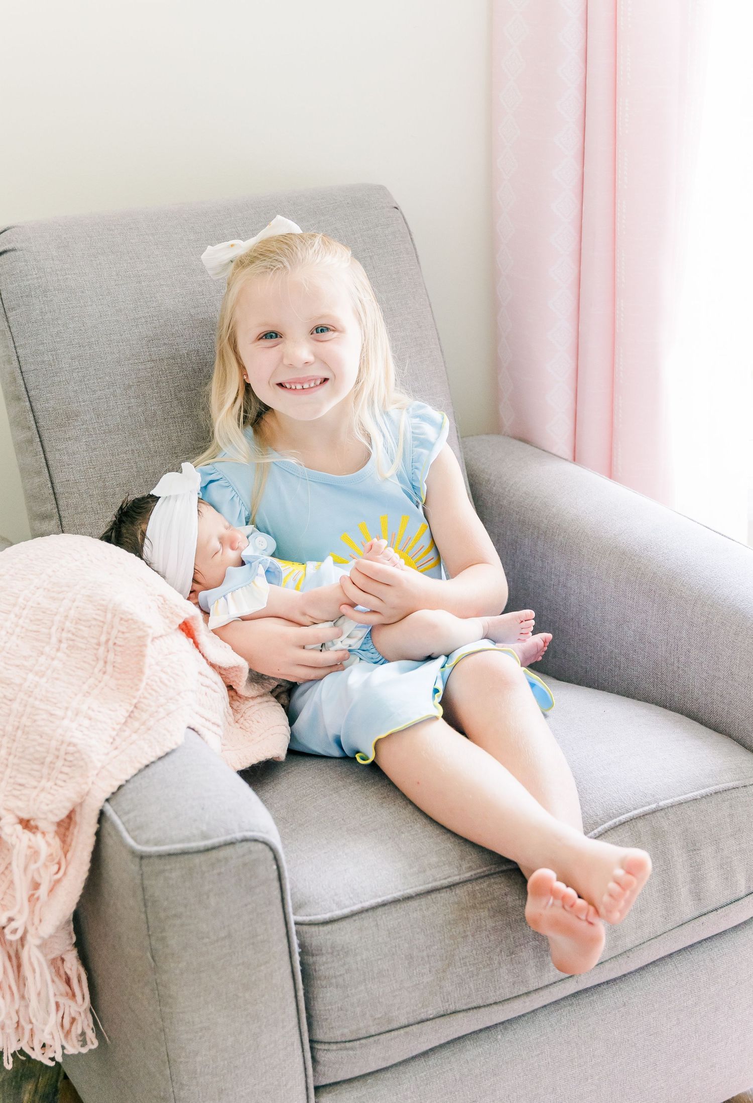Young child in light blue outfit sits happily on a gray armchair with a plush toy.
