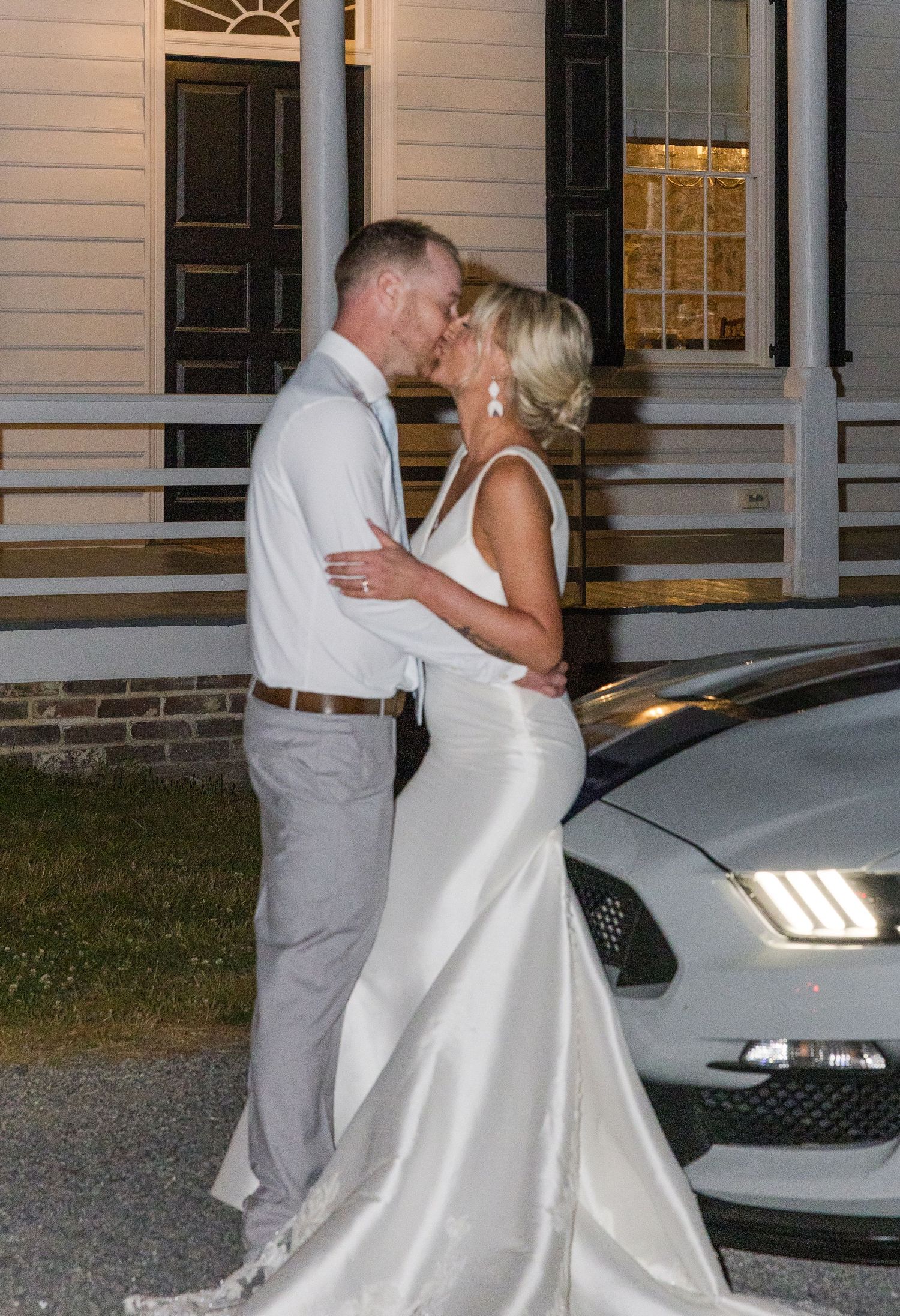 Bride and groom share a romantic kiss beside a Ford Mustang at dusk outside a historic building.