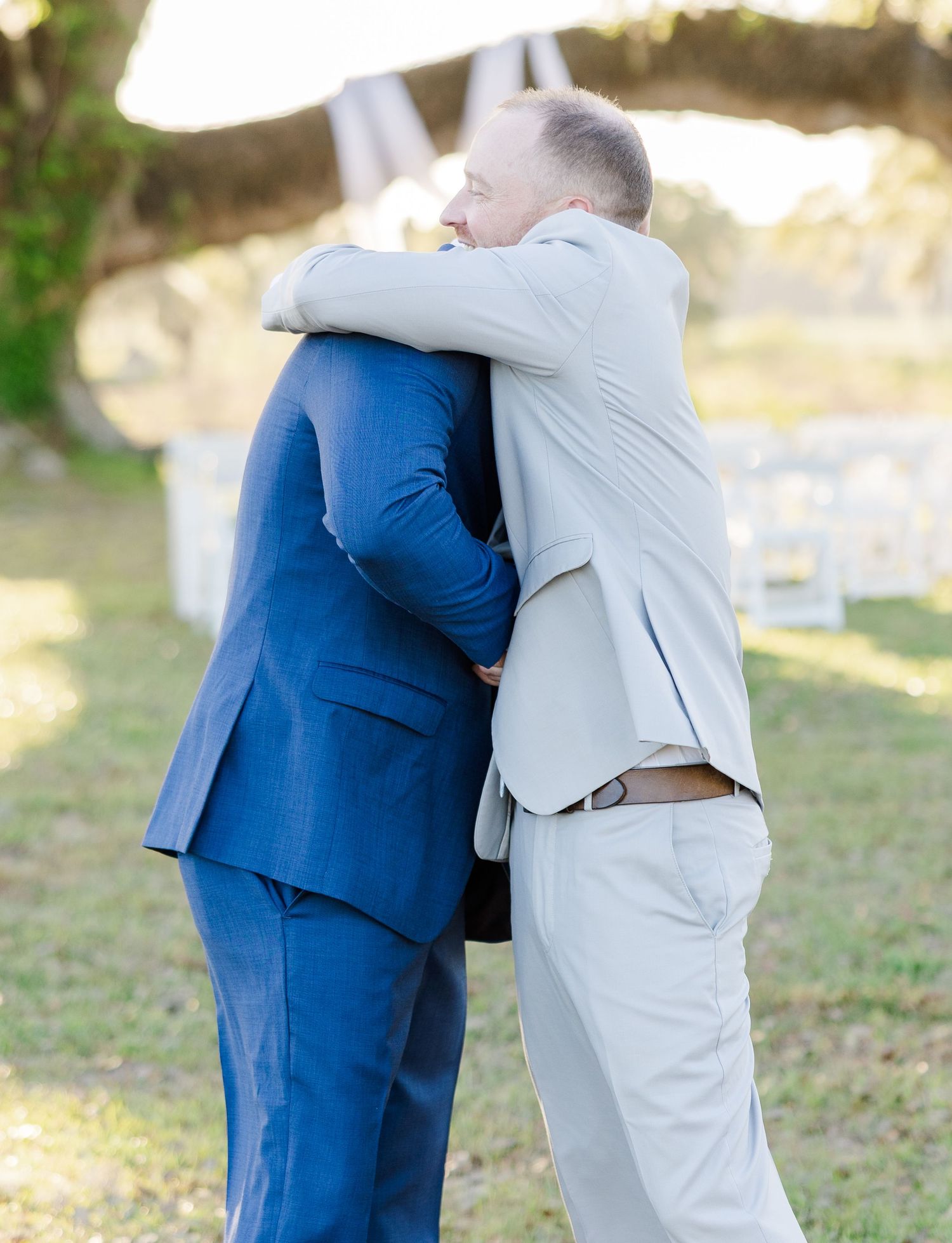 Two men in suits share an emotional hug at an outdoor wedding ceremony beneath a large oak tree.