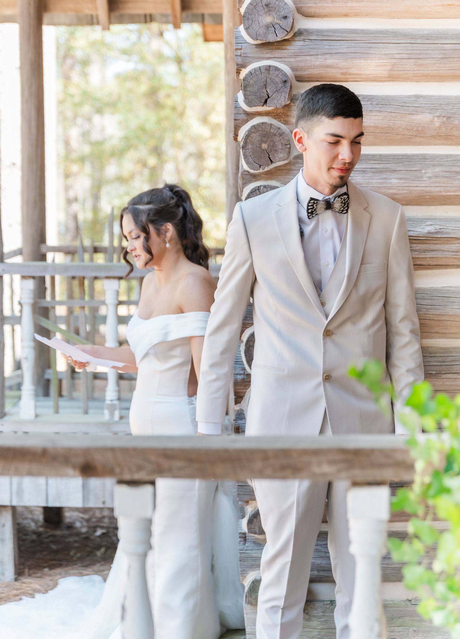 Bride in off-shoulder gown reads vows while groom in tan suit with bow tie stands against log cabin wall.
