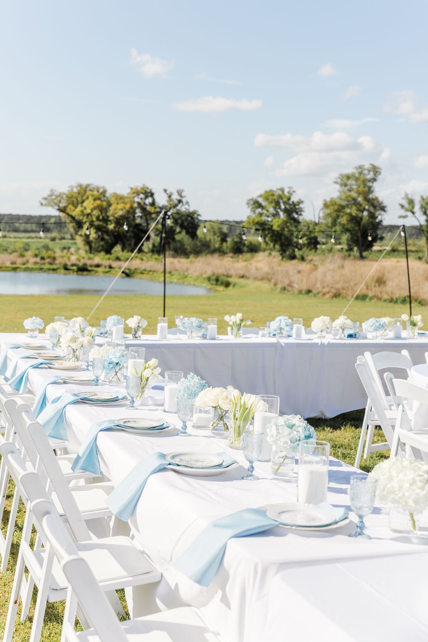 Elegant outdoor wedding reception tables with blue napkins, white florals, and waterfront view.