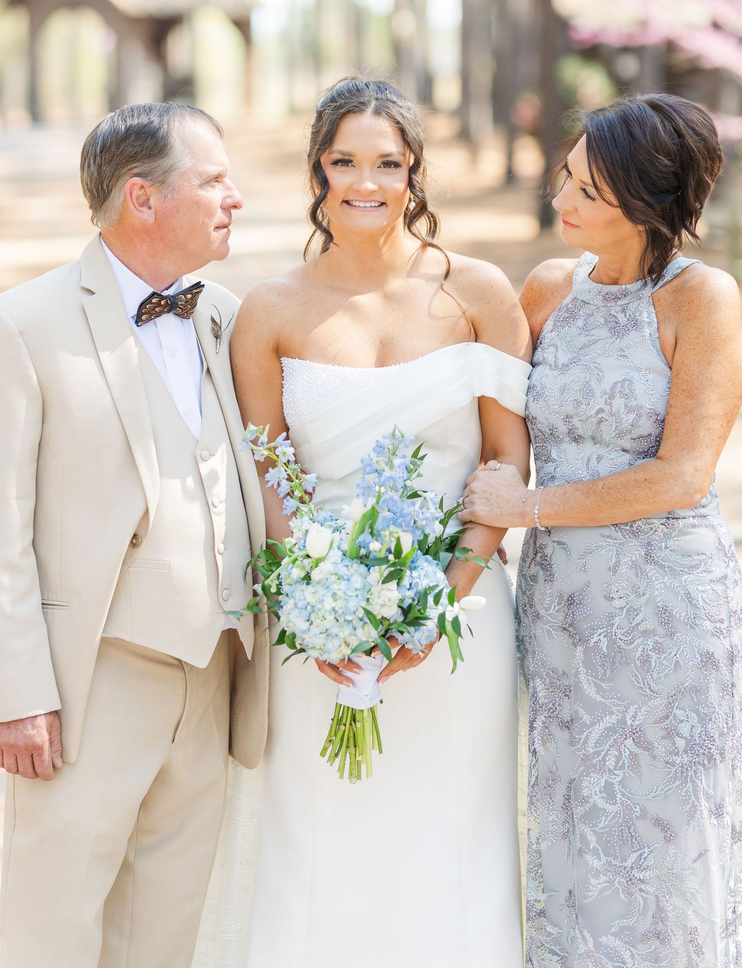 Bride with blue and white bouquet posing with parents in cream suit and floral dress at outdoor wedding.