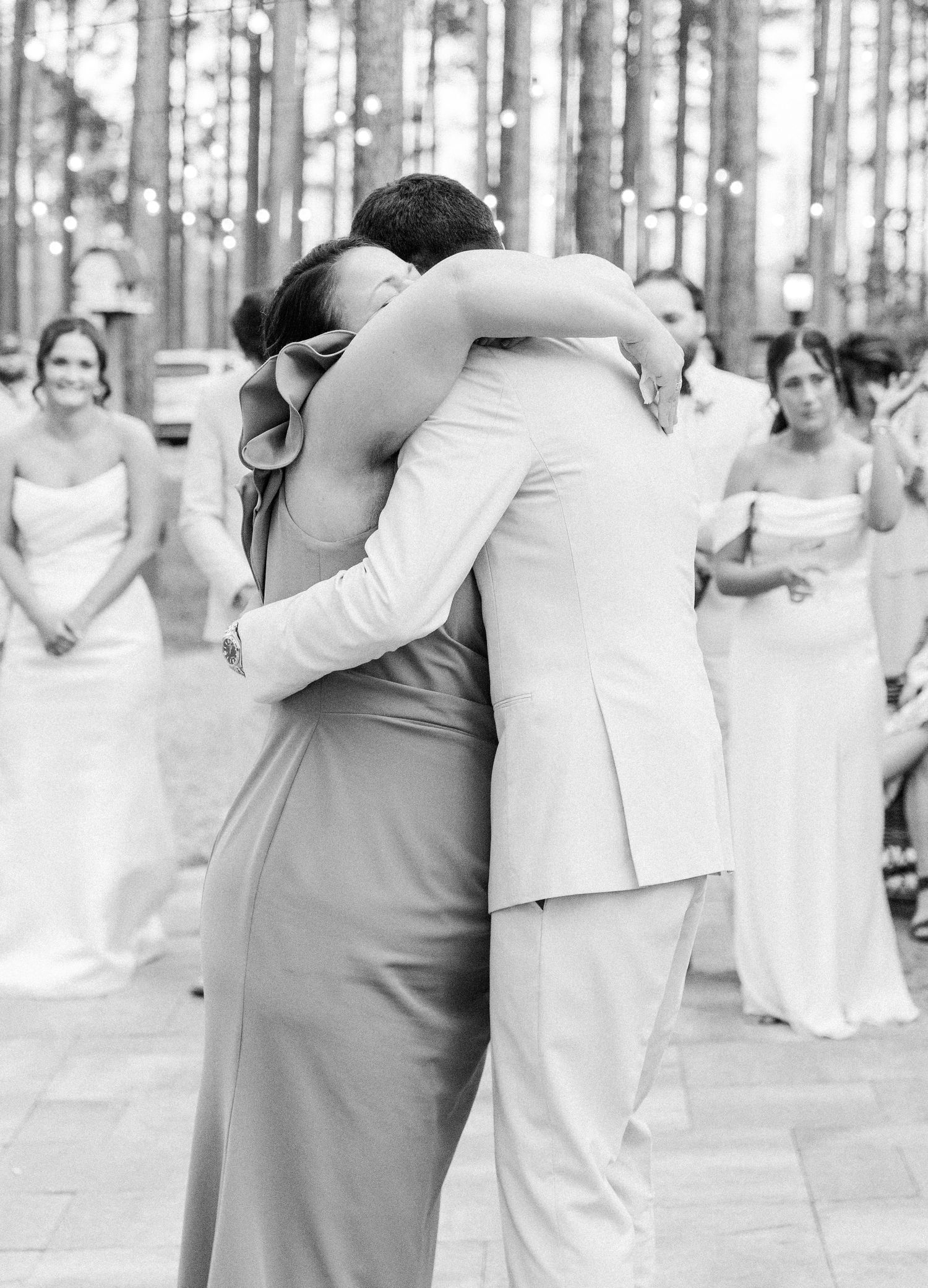 Black and white photo of a couple sharing an emotional hug at their outdoor wedding ceremony surrounded by guests.